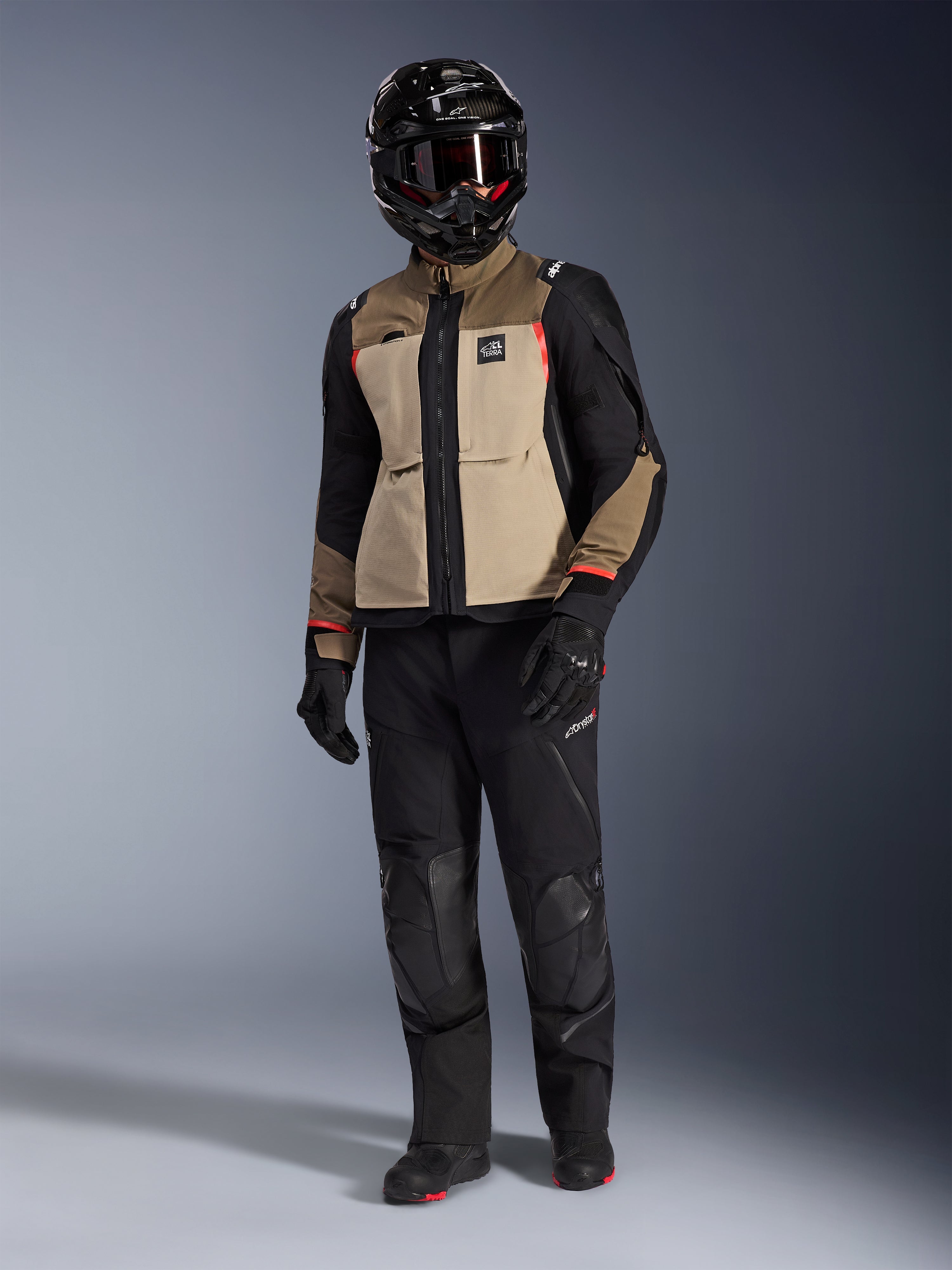 Kurtka Alpinestars Andes Pro Drystar® XF w kolorze Walnut Aluminum, motocyklowa kurtka adventure noszona z pasującymi czarnymi spodniami, czarnym kaskiem adventure z goglami i czarnymi butami, z beżowymi i czarnymi panelami oraz czerwonymi akcentami.