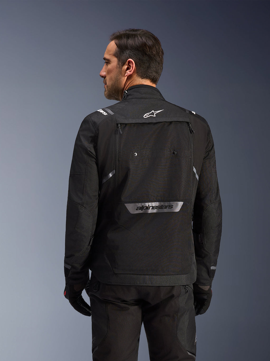 Rider from behind wearing Alpinestars Cusco Drystar® Jacket in Black, textile adventure touring jacket with large rear storage pocket and reflective branding, paired with matching black pants and gloves