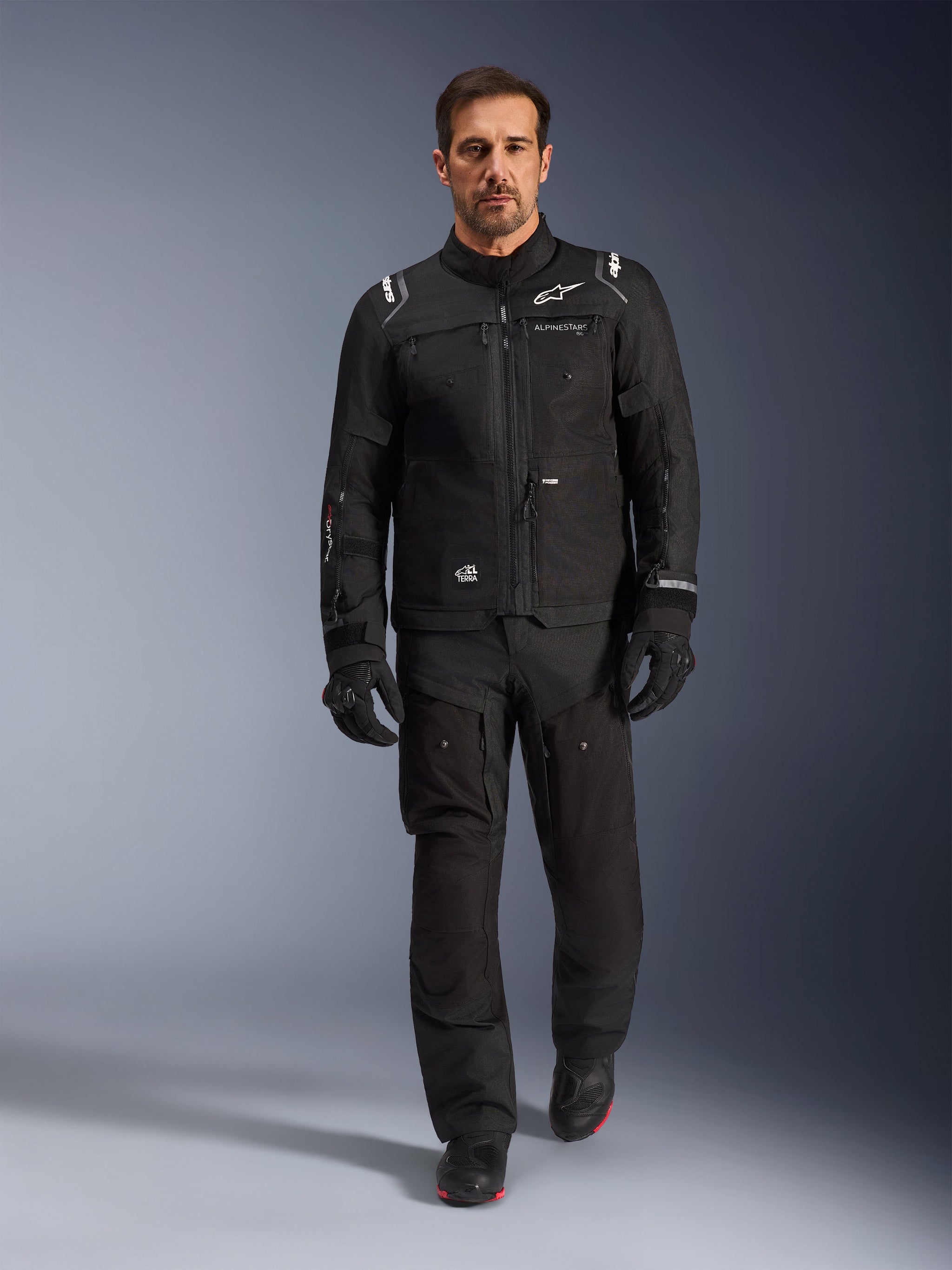 Motocyklista ubrany w czarną kurtkę Alpinestars Cusco Drystar®, w zestawie z dopasowanymi czarnymi spodniami technicznymi i rękawicami, studyjne ujęcie całej sylwetki, odzież motocyklowa adventure touring, widoczny branding Alpinestars i konstrukcja z wieloma kieszeniami.