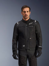 Alpinestars Cusco Drystar® Jacket, czarna kurtka motocyklowa, prezentowana przez modela w dopasowanych czarnych rękawicach i spodniach, techniczna konstrukcja tekstylna z białymi logotypami Alpinestars, zaprojektowana do turystyki szosowej typu adventure