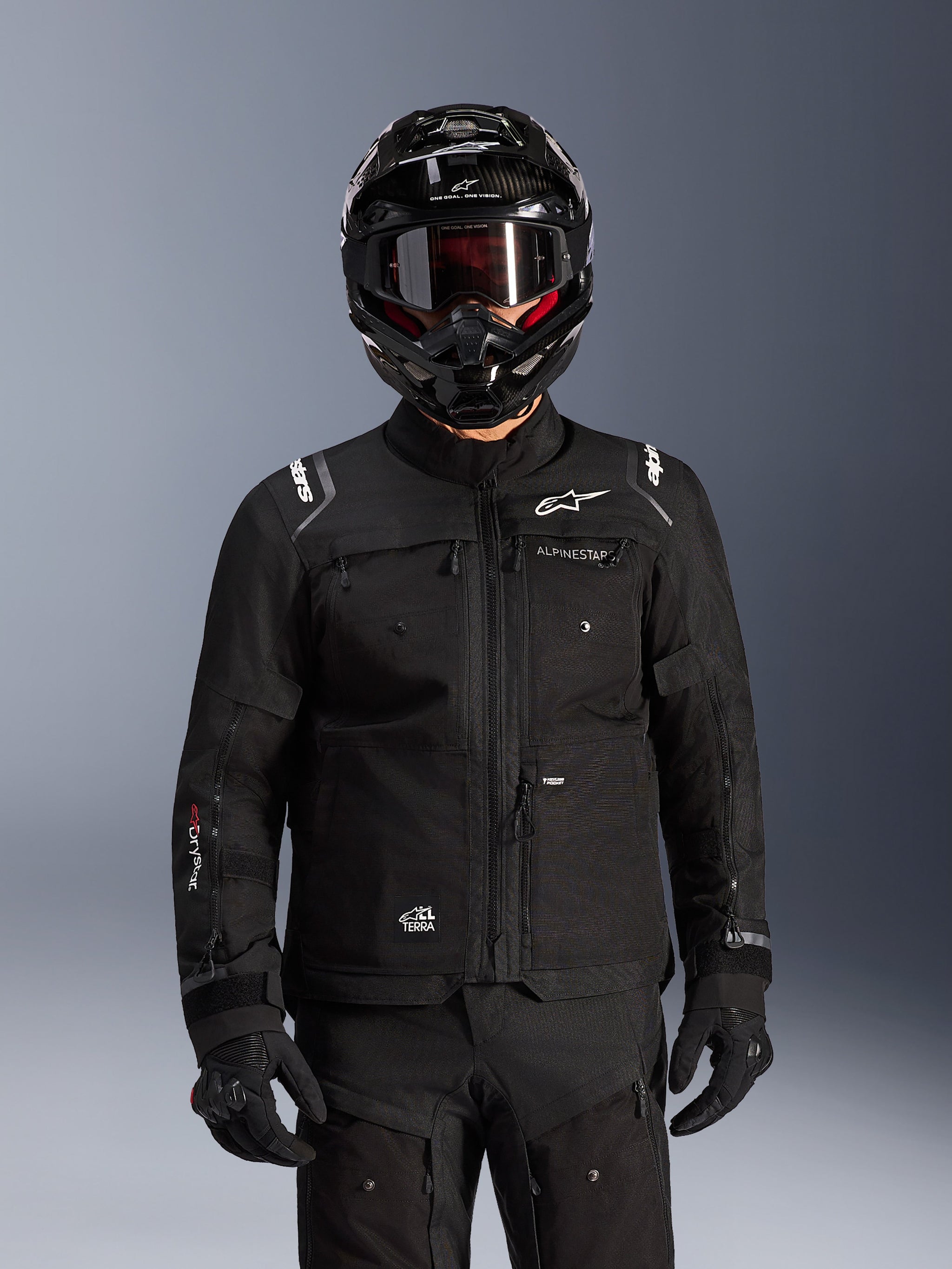 Kurtka Alpinestars Cusco Drystar®, czarna motocyklowa kurtka turystyczna adventure, noszona z czarnym kaskiem adventure i ciemnymi goglami, wyposażona w liczne kieszenie użytkowe, logo Alpinestars na klatce piersiowej i logo Drystar® na rękawie, element kompletnego stroju wyprawowego.