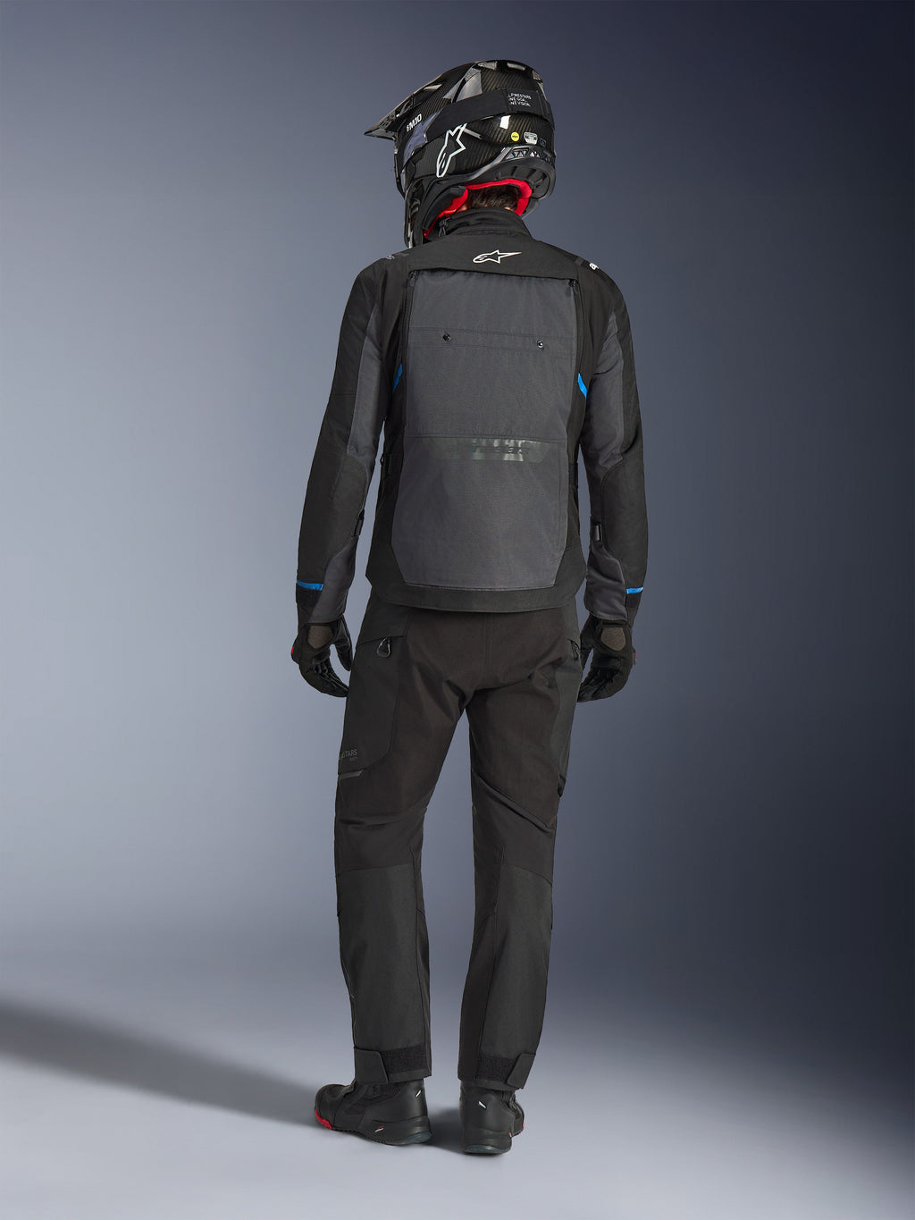 Kurtka Alpinestars Cusco Drystar®, czarna i ciemnoszara, widok z tyłu motocyklisty w pełnym stroju adventure touring, duży szary panel na plecach z logo Alpinestars, niebieskie akcenty na rękawach, noszona z pasującymi czarnymi spodniami i kaskiem.