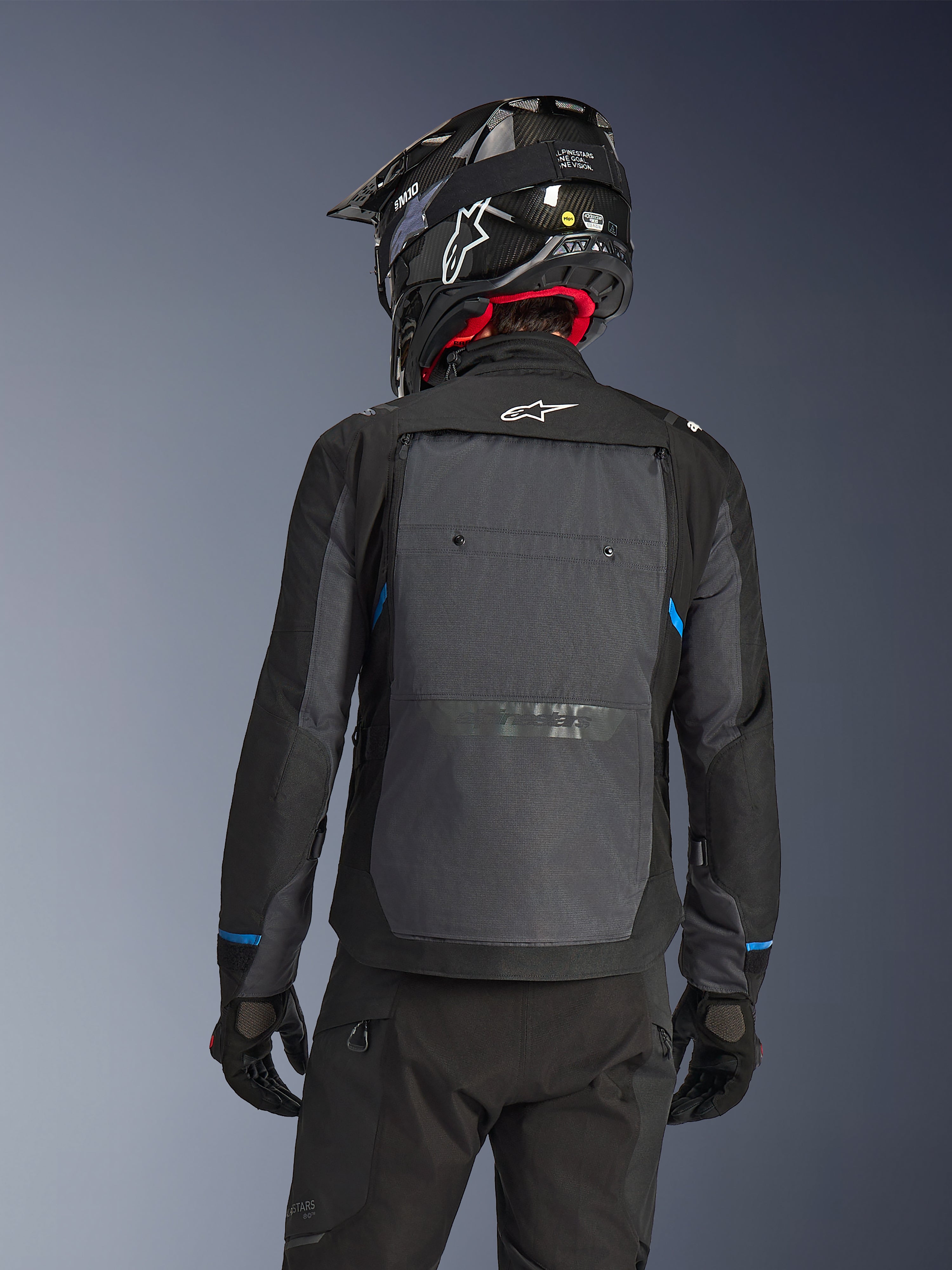 Motocyklista widziany od tyłu w kurtce Alpinestars Cusco Drystar® w kolorze czarnym i ciemnoszarym, z dużym szarym panelem wentylacyjnym na plecach i białym logo, w czarnym kasku terenowym i czarnym stroju do jazdy adventure touring.