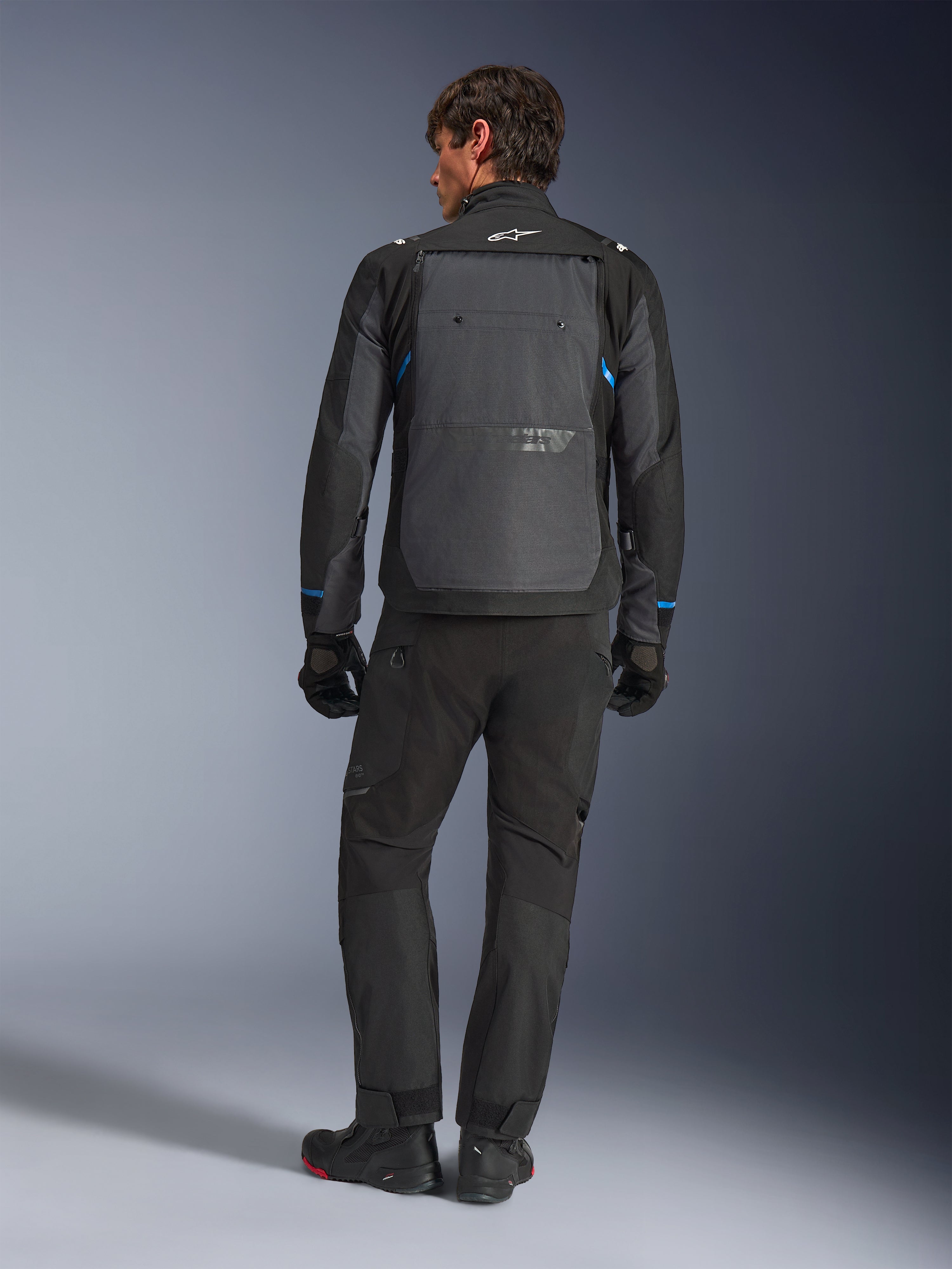Alpinestars Cusco Drystar® Jacket, czarno-ciemnoszara z niebieskimi akcentami, widok z tyłu, noszona z dopasowanymi czarnymi spodniami adventure i rękawicami, z dużym szarym panelem na plecach i białym logo na kołnierzu, zaprojektowana do turystyki szosowej i adventure touring