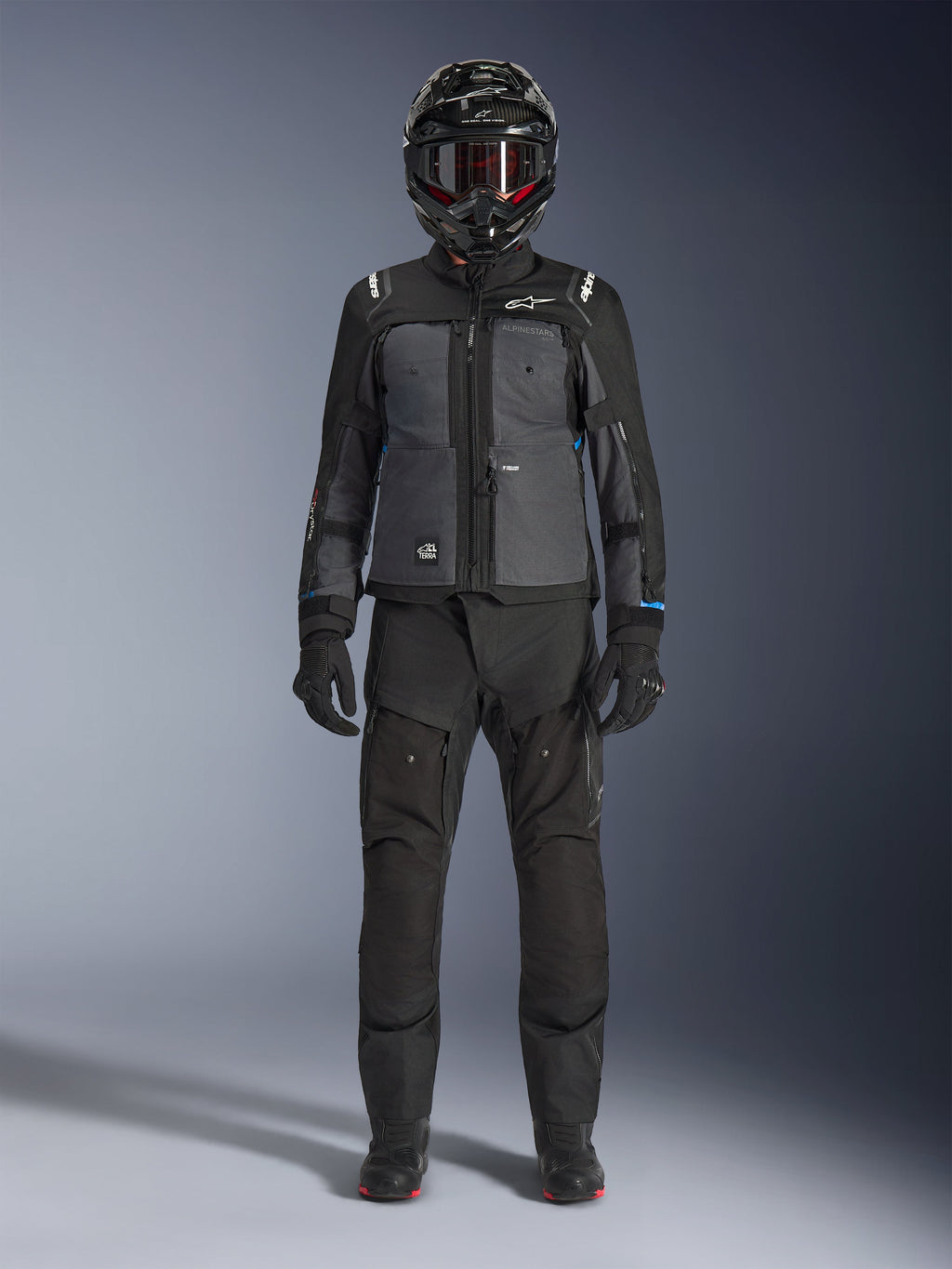 Kurtka Alpinestars Cusco Drystar®, czarno-ciemnoszara kurtka adventure touring, noszona z dopasowanymi czarnymi spodniami, rękawicami i kaskiem integralnym, widok na całą sylwetkę od przodu, ochronna odzież motocyklowa
