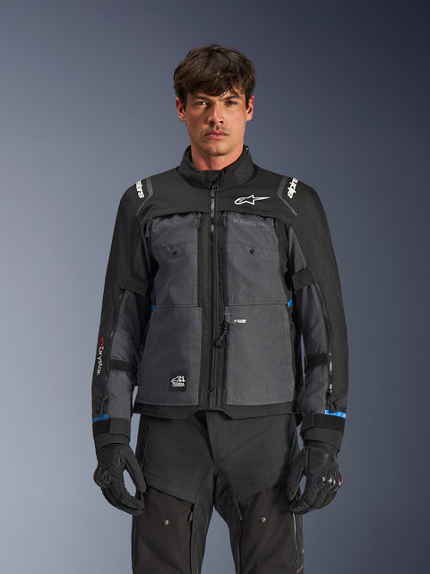 Kurtka Alpinestars Cusco Drystar®, motocyklowa kurtka turystyczna, czarna i ciemnoszara, widok z przodu na modelu, logo Alpinestars na klatce piersiowej i ramionach, branding Drystar® na rękawach, techniczna konstrukcja z wieloma kieszeniami i zamkami wentylacyjnymi, w połączeniu z czarną odzieżą motocyklową