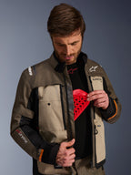 Kurtka Alpinestars Cusco Drystar®, Walnut Aluminum Black, noszona przez mężczyznę prezentującego kieszeń na ochraniacz klatki piersiowej z czerwoną wkładką ochronną, tekstylna kurtka motocyklowa z otworami wentylacyjnymi na zamek i brandingiem adventure touring.