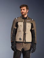 Kurtka Alpinestars Cusco Drystar®, przygodowa kurtka motocyklowa, kolor Walnut Aluminum Black, prezentowana przez modela w czarnych rękawicach, z beżowymi panelami na klatce piersiowej i czarnymi rękawami, logo Alpinestars i naszywka All Terra, ochronna odzież turystyczna na każdą pogodę