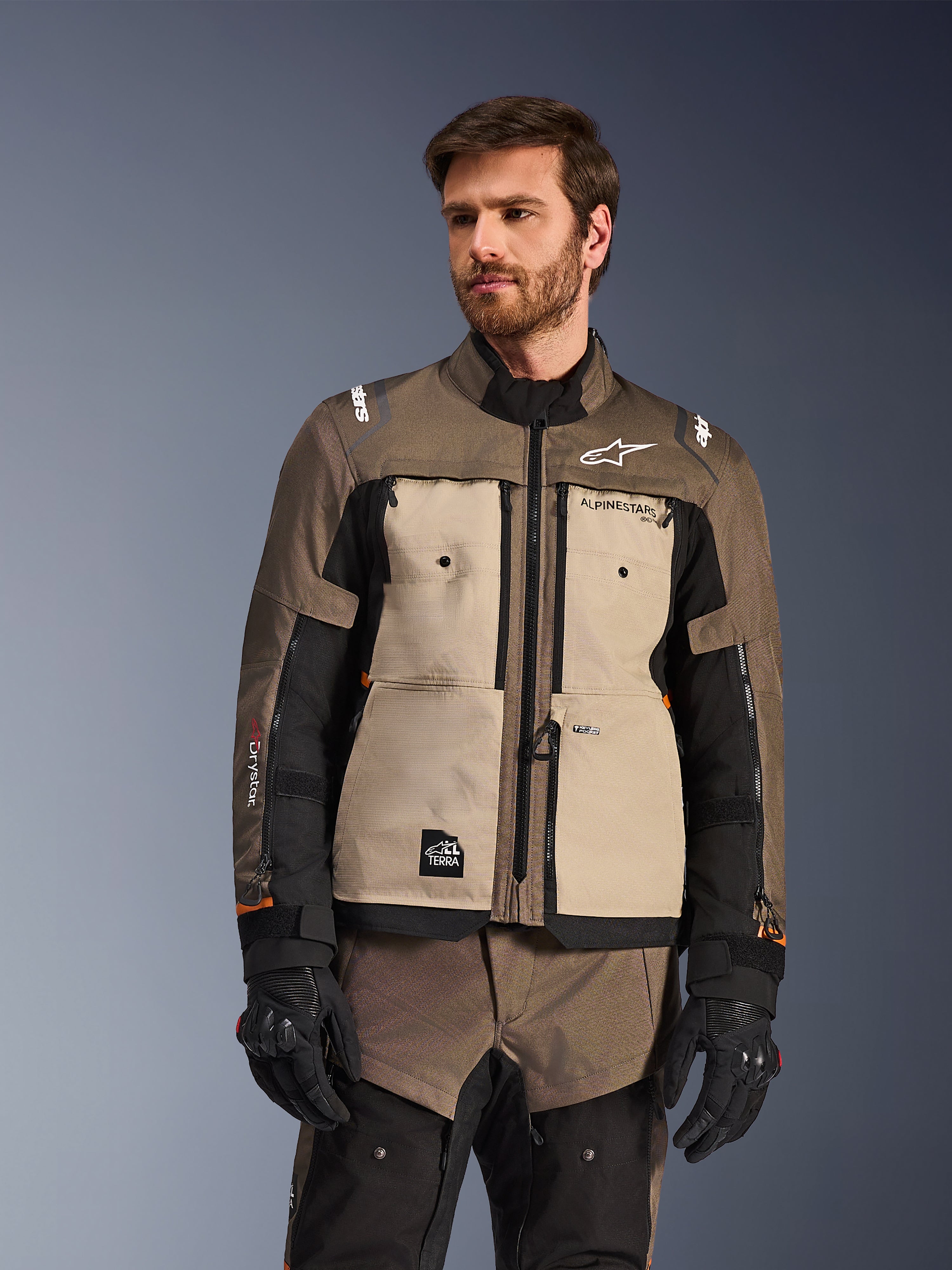 Kurtka Alpinestars Cusco Drystar®, przygodowa kurtka motocyklowa, kolor Walnut Aluminum Black, prezentowana przez modela w czarnych rękawicach, z beżowymi panelami na klatce piersiowej i czarnymi rękawami, logo Alpinestars i naszywka All Terra, ochronna odzież turystyczna na każdą pogodę