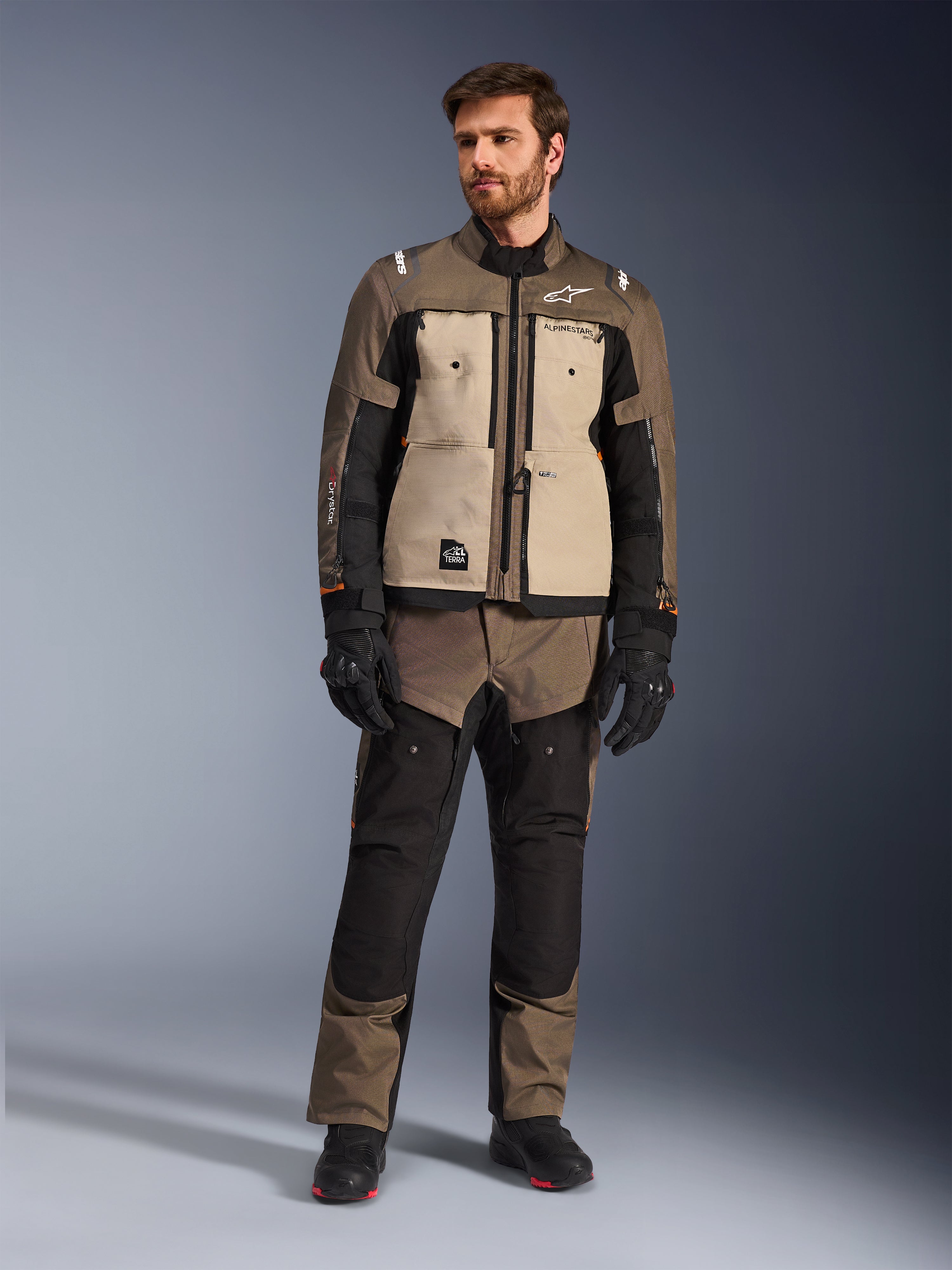 Kurtka Alpinestars Cusco Drystar® w kolorze Walnut Aluminum Black, noszona przez motocyklistę z pasującymi spodniami adventure touring, czarnymi rękawicami i butami, wyposażona w beżowe panele na klatce piersiowej, czarne rękawy z subtelnymi pomarańczowymi akcentami i funkcjonalne zamki wentylacyjne do drogowej jazdy motocyklowej.