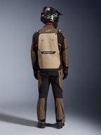 Motocyklista widziany od tyłu w kurtce Alpinestars Cusco Drystar® w kolorze Walnut Aluminum Black, z dużą tylną kieszenią w kolorze aluminium i pomarańczowymi akcentami, pasujące spodnie adventure, czarny kask off-road i rękawice, wytrzymała tekstylna odzież motocyklowa turystyczna.