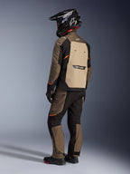 Kurtka Alpinestars Cusco Drystar®, kurtka turystyczna adventure, Walnut Aluminum Black, widok z tyłu prezentujący beżowy panel tylny i pomarańczowe akcenty, noszona z pasującymi spodniami i czarnym kaskiem z włókna węglowego