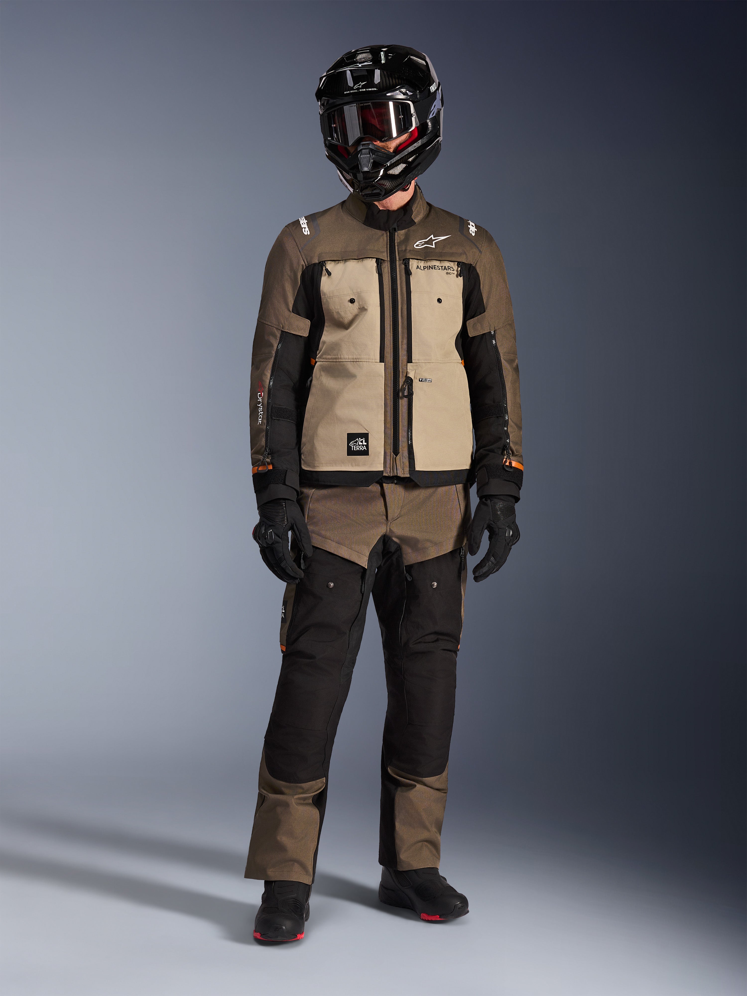 Kurtka Alpinestars Cusco Drystar®, Walnut Aluminum Black, motocyklowa kurtka turystyczna adventure noszona z pasującymi spodniami i czarnym kaskiem, z beżowymi panelami na klatce piersiowej i brązowymi rękawami ripstop, techniczna odzież motocyklowa na każdą pogodę