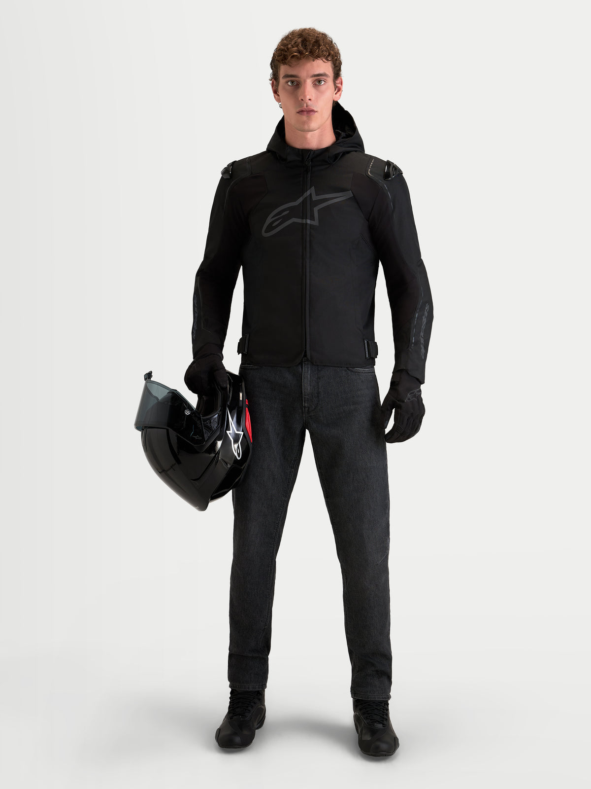 Alpinestars Zaca V2 Waterproof, męska czarna kurtka motocyklowa z kapturem i ochraniaczami ramion, noszona przez motocyklistę trzymającego kask.