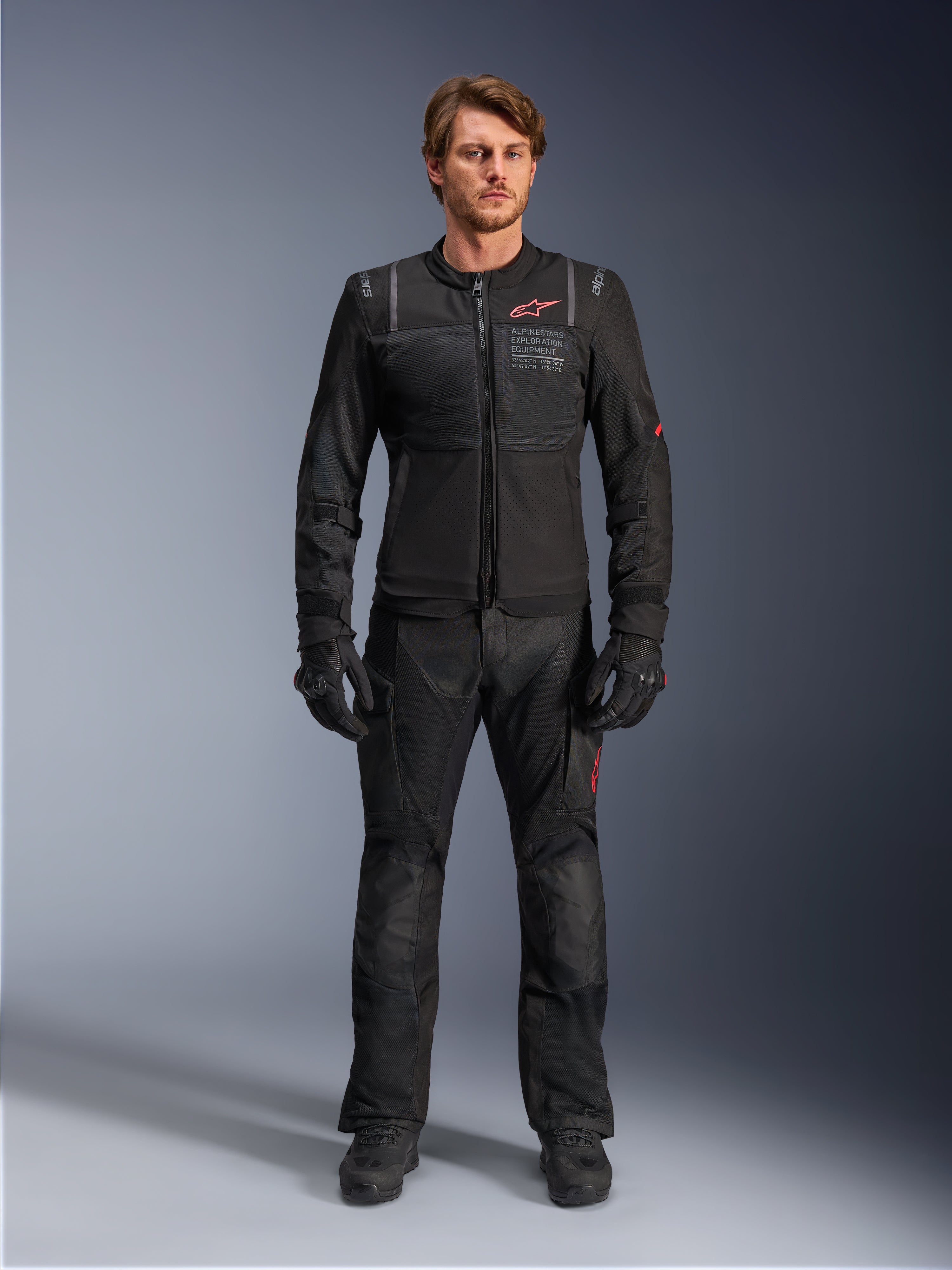 Męski model w kurtce Alpinestars ST-2 Air, czarna kurtka motocyklowa z panelami z siatki i czerwonym logo, napis „Alpinestars Exploration Equipment” na klatce piersiowej, w zestawieniu z czarnymi technicznymi spodniami i rękawicami, nowoczesny strój adventure touring