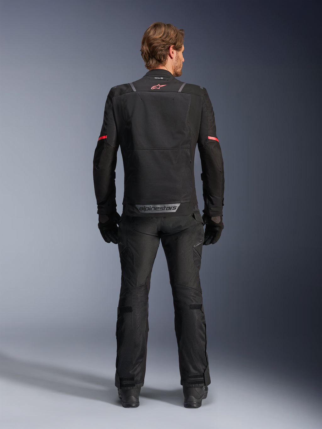 Kurtka Alpinestars ST-2 Air, czarna, widok od tyłu motocyklisty w kurtce motocyklowej i pasujących czarnych spodniach, panele wentylacyjne z siatki, czerwone logo w górnej części pleców, szary napis Alpinestars na dolnym brzegu pleców