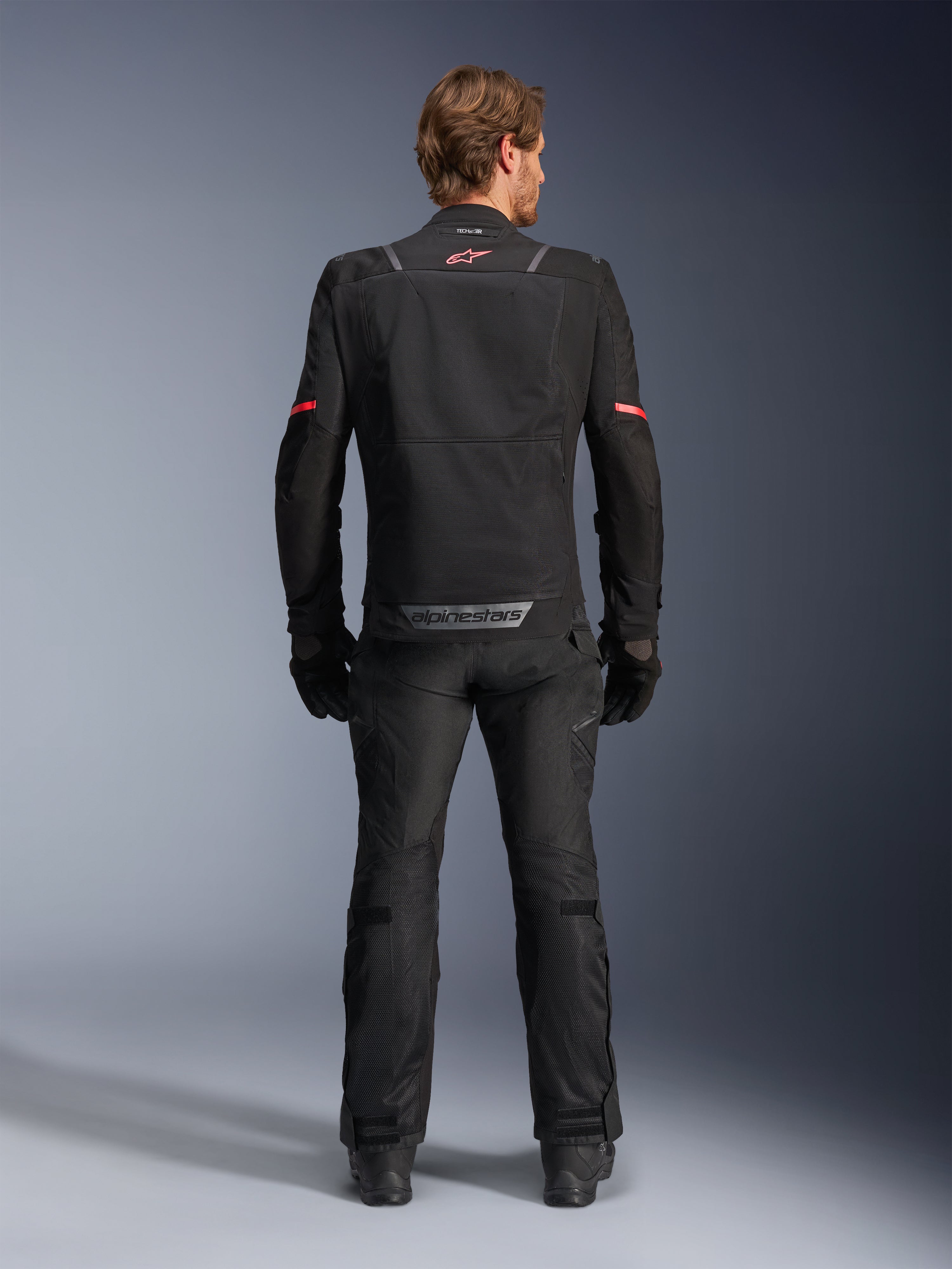 Kurtka Alpinestars ST-2 Air, czarna, widok od tyłu motocyklisty w kurtce motocyklowej i pasujących czarnych spodniach, panele wentylacyjne z siatki, czerwone logo w górnej części pleców, szary napis Alpinestars na dolnym brzegu pleców
