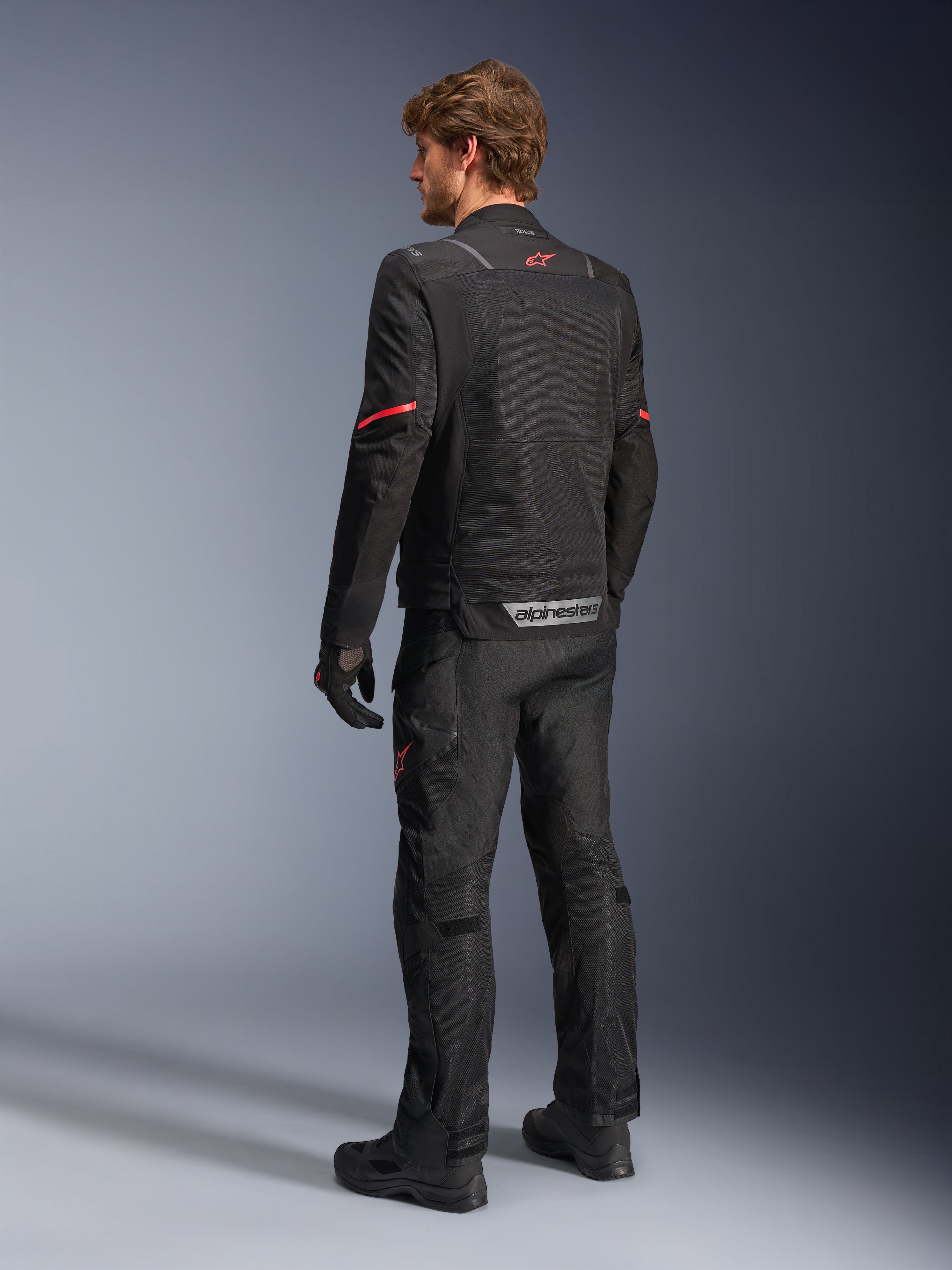 Alpinestars ST-2 Air Jacket, czarna kurtka motocyklowa, widok z tyłu na modelu z czerwonym logo gwiazdy w górnej części pleców i odblaskowym brandingiem Alpinestars w pasie, w zestawieniu z pasującymi czarnymi technicznymi spodniami i rękawicami.