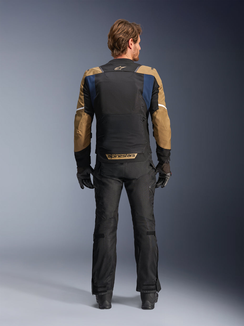 Kurtka Alpinestars ST-2 Air w kolorach Deep Blue, Dark Khaki i czarnym, widok od tyłu na modelu pokazujący duży siatkowy panel na plecach oraz rękawy w kolorze khaki z białymi paskami, w zestawieniu z czarnymi technicznymi spodniami motocyklowymi i rękawicami do turystyki adventure.
