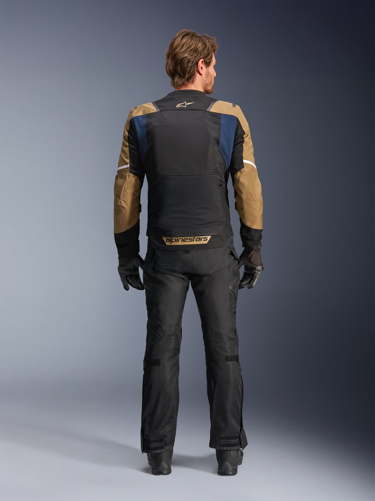 Kurtka Alpinestars ST-2 Air w kolorach Deep Blue, Dark Khaki i czarnym, widok od tyłu na modelu pokazujący duży siatkowy panel na plecach oraz rękawy w kolorze khaki z białymi paskami, w zestawieniu z czarnymi technicznymi spodniami motocyklowymi i rękawicami do turystyki adventure.