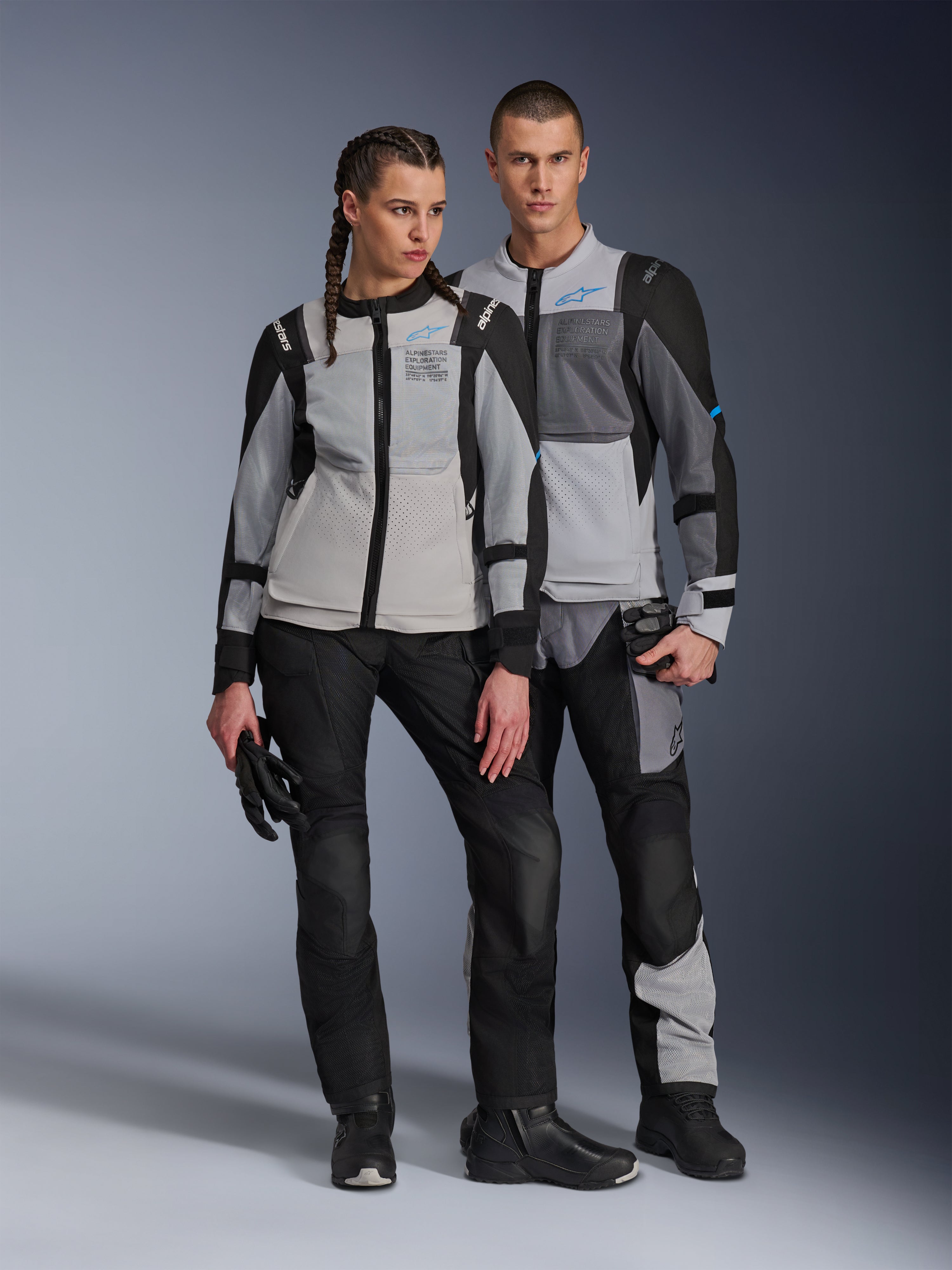 Modele (kobieta i mężczyzna) ubrani w kurtki Alpinestars ST-2 Air w kolorach Ice Gray, Dark Gray i Black, techniczna letnia turystyczna odzież motocyklowa z dużymi panelami z siatki i brandingiem exploration equipment, w zestawie z czarnymi spodniami, rękawicami i butami.