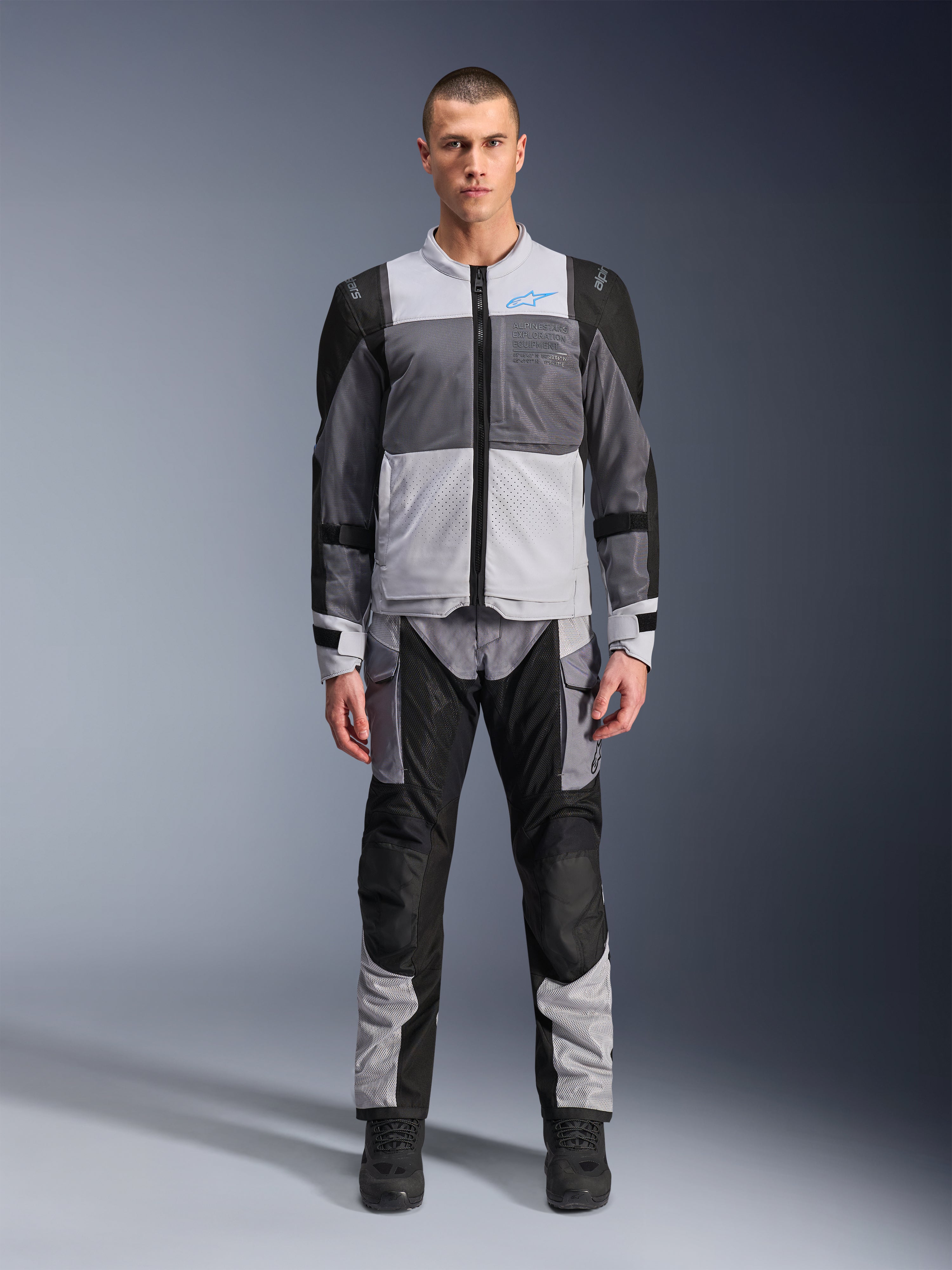 Kurtka Alpinestars ST-2 Air, motocyklowa kurtka turystyczna, kolory lodowy szary, ciemnoszary i czarny, prezentowana na modelu z dopasowanymi spodniami adventure, duże panele z siatki, niebieskie logo Alpinestars na klatce piersiowej, branding Alpinestars Exploration Equipment