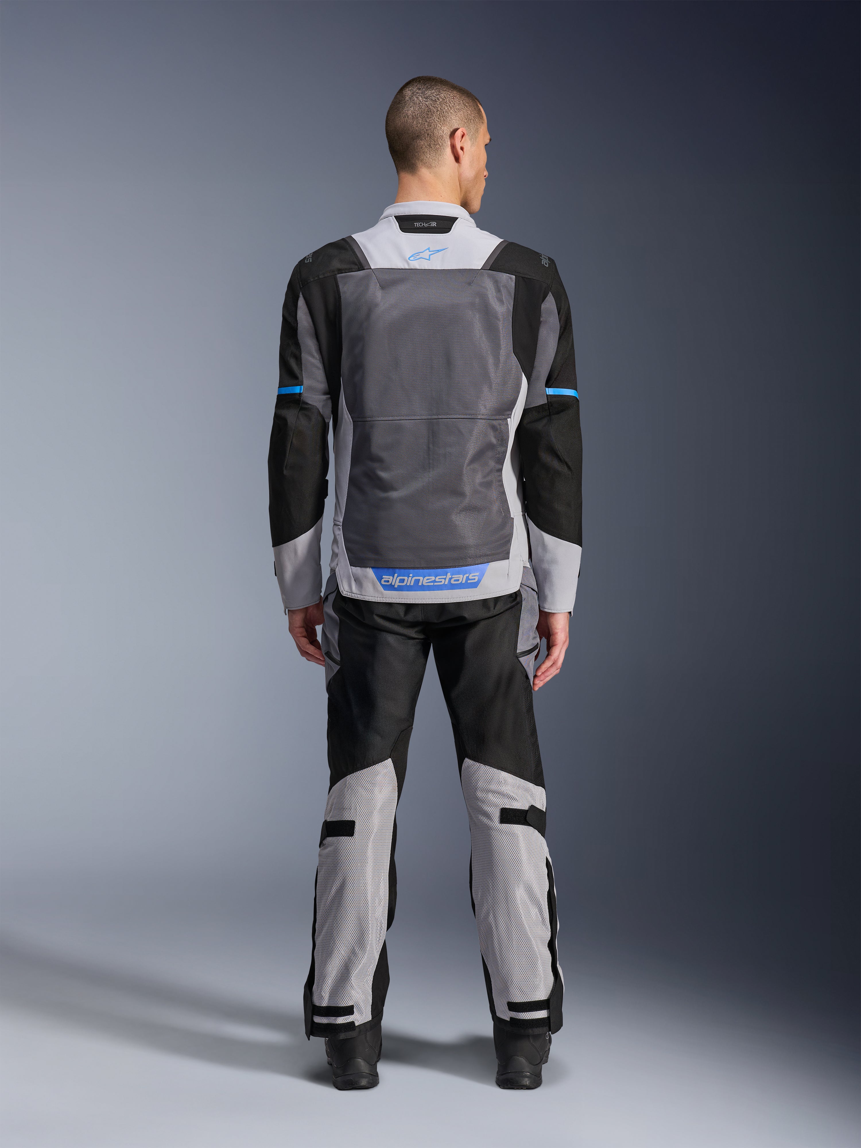 Alpinestars ST-2 Air Jacket, kolor Ice Gray Dark Gray Black, widok z tyłu na modelu, duże siatkowe panele wentylacyjne, niebieskie akcenty i logo Alpinestars, w zestawie z pasującymi spodniami motocyklowymi do adventure touring.