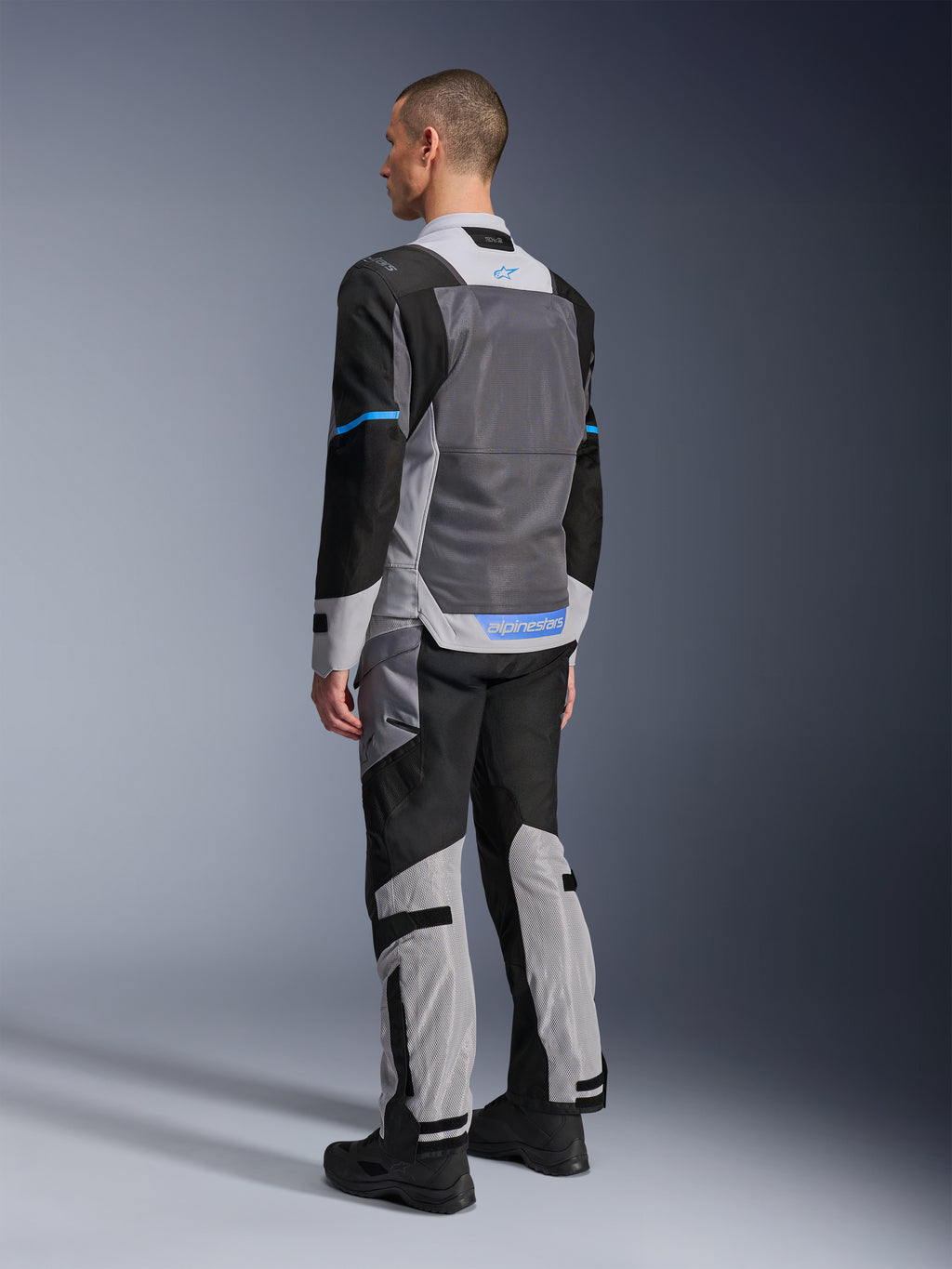 Męski model widoczny od tyłu w kurtce Alpinestars ST-2 Air w kolorach Ice Gray, Dark Gray i czarnym, z dużym szarym panelem z siatki na plecach, niebieskimi akcentami logo i dopasowanymi spodniami motocyklowymi do letniej turystyki.