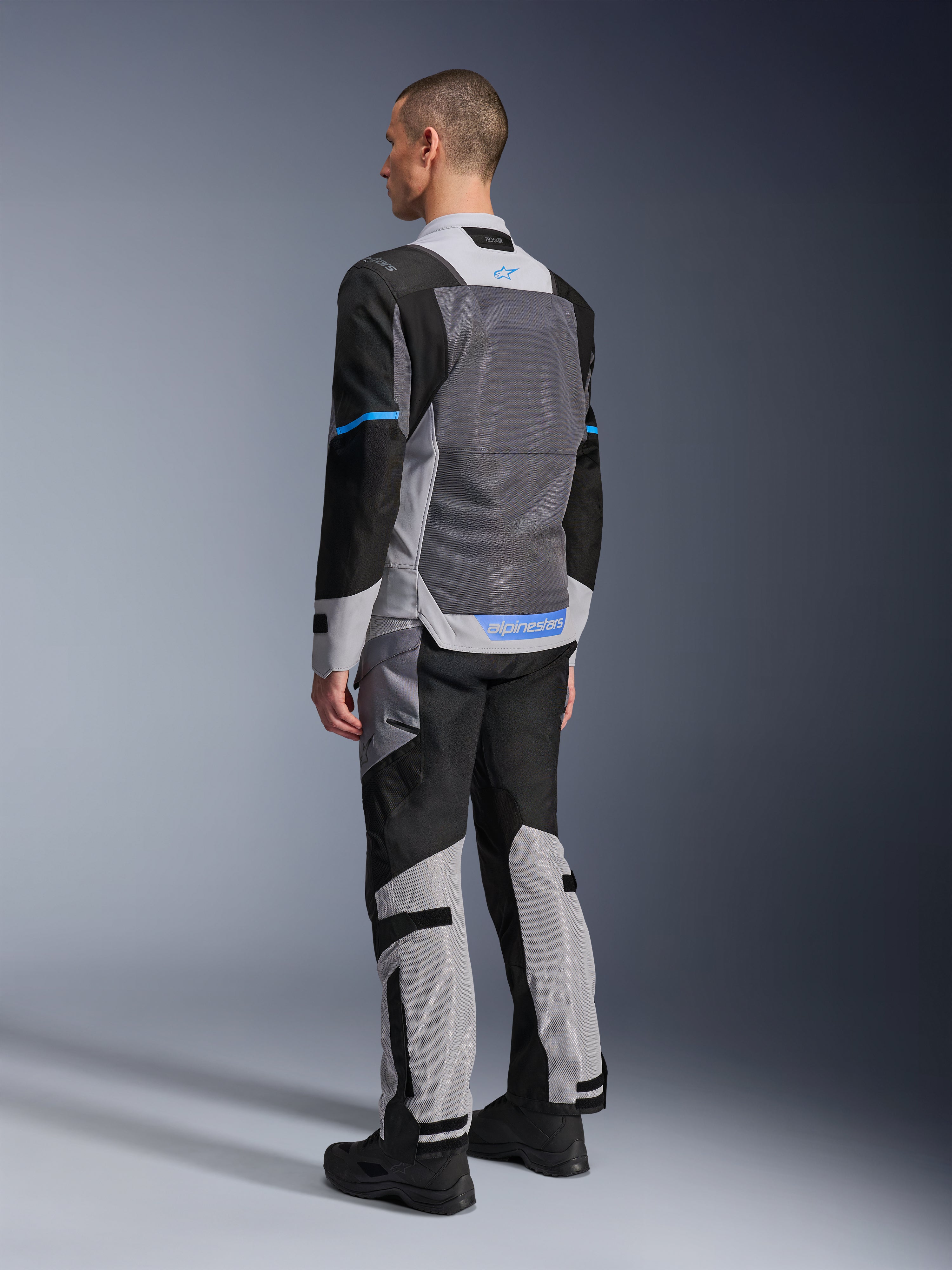Męski model widoczny od tyłu w kurtce Alpinestars ST-2 Air w kolorach Ice Gray, Dark Gray i czarnym, z dużym szarym panelem z siatki na plecach, niebieskimi akcentami logo i dopasowanymi spodniami motocyklowymi do letniej turystyki.