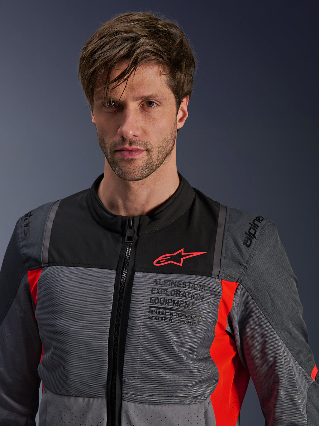 Kurtka Alpinestars ST-2 Air, ciemnoszara i jasnoszara z jaskrawoczerwonymi akcentami, letnia turystyczna kurtka motocyklowa prezentowana przez modela, siatkowe panele wentylacyjne, czerwone logo Astars i napis „Exploration Equipment” na klatce piersiowej