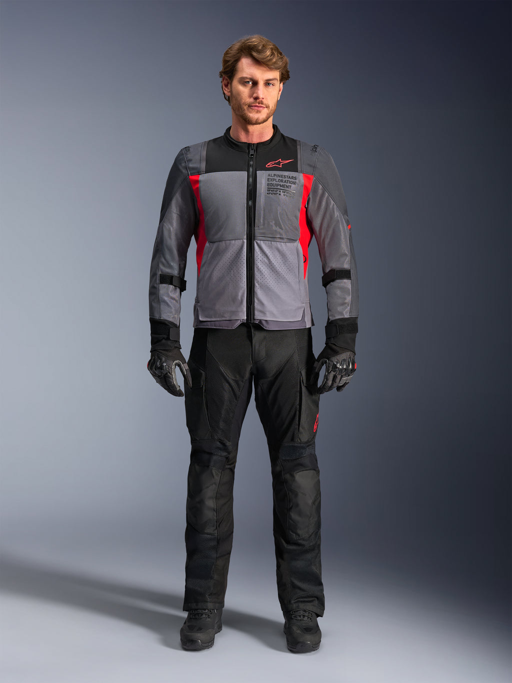 Kurtka Alpinestars ST-2 Air w kolorze ciemnoszarym i jaskrawoczerwonym, na modelu z czarnymi spodniami motocyklowymi i rękawicami, duże panele z siatki dla przepływu powietrza, nowoczesne oznakowanie exploration equipment
