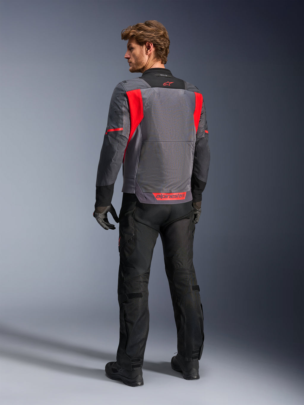 Mężczyzna widoczny od tyłu w kurtce Alpinestars St-2 Air w kolorach Deep Gray, Dark Gray i Bright Red, prezentujący duży siatkowy panel wentylacyjny na plecach i czerwone oznaczenia marki, zestawiony z czarnymi spodniami motocyklowymi i szarymi rękawicami.