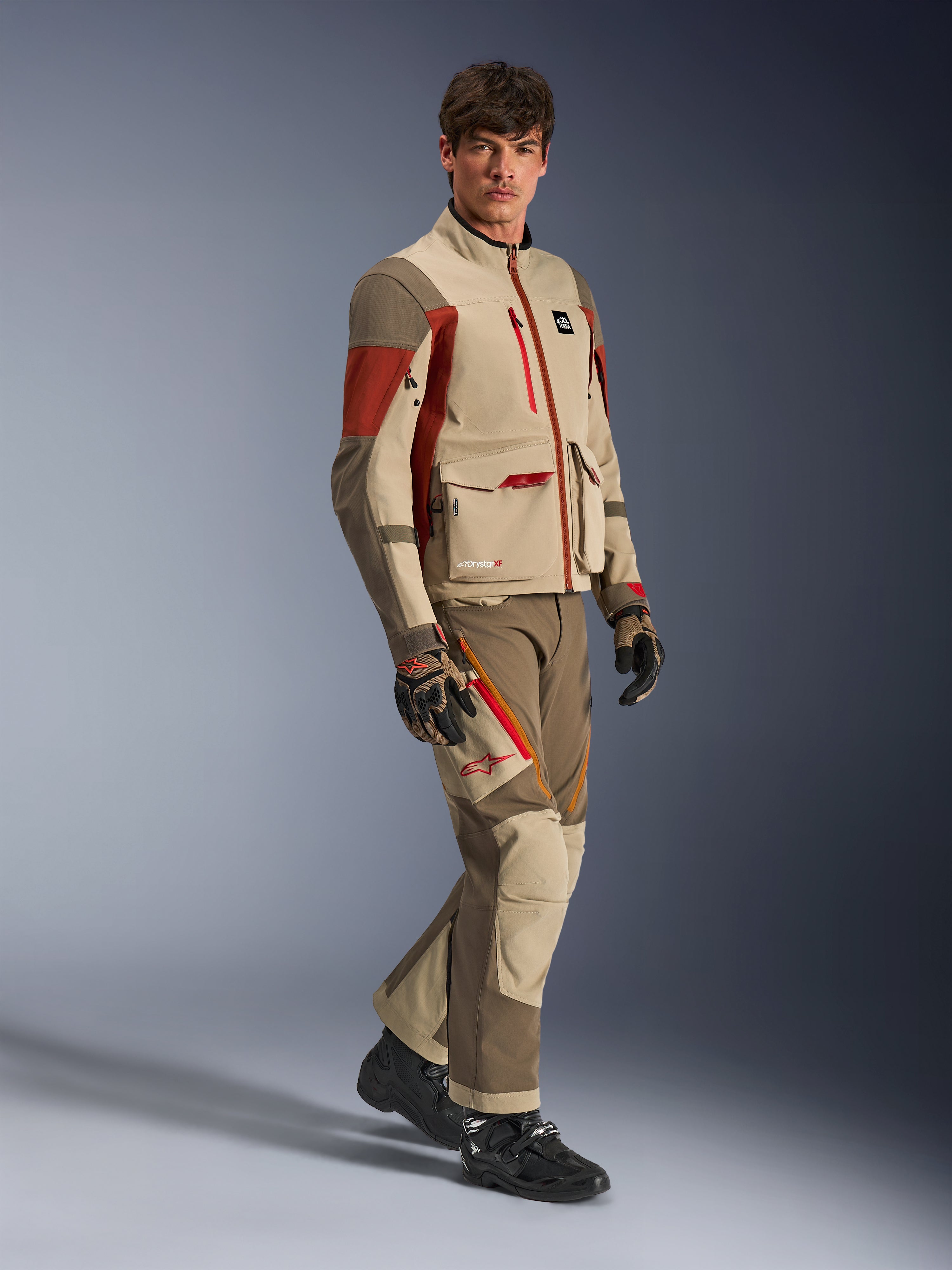 Męski model w kurtce Alpinestars Maxdura Drystar®XF Laminated Armored Shell w kolorze Aluminum Walnut Rust Brown, pasujące spodnie, czarno-brązowe rękawice i czarne buty, wielowarstwowa techniczna konstrukcja z kieszeniami cargo i czerwonymi detalami zamków, dopasowany strój do turystyki przygodowej