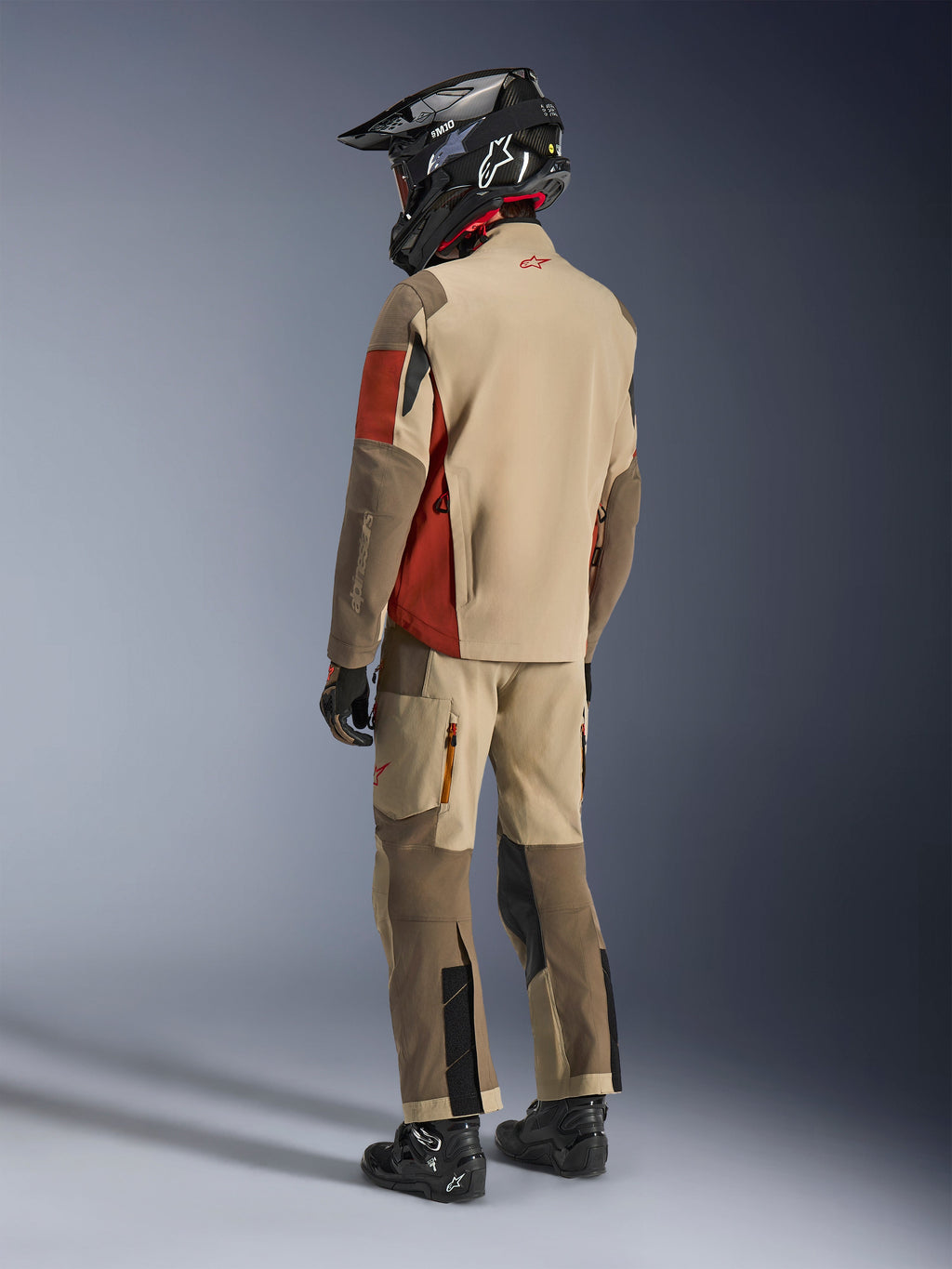 Motocyklista adventure widoczny z tyłu, ubrany w kurtkę Alpinestars Maxdura Drystar®XF Laminated Armored Shell w kolorze Aluminum Walnut Rust Brown, w zestawie z pasującymi spodniami motocyklowymi, czarnym kaskiem off-road i czarnymi butami; kompletny techniczny strój turystyczny.