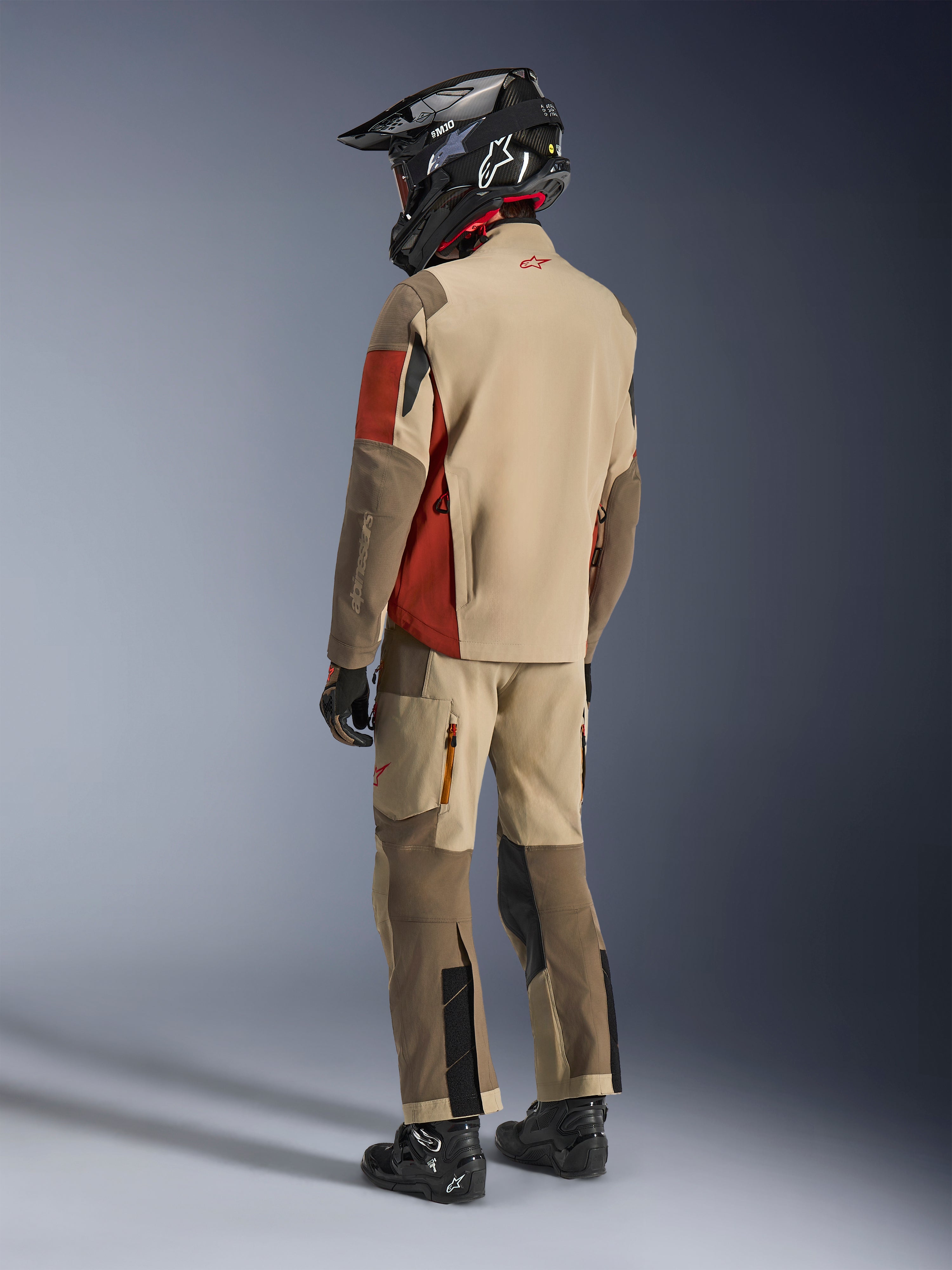 Motocyklista adventure widoczny z tyłu, ubrany w kurtkę Alpinestars Maxdura Drystar®XF Laminated Armored Shell w kolorze Aluminum Walnut Rust Brown, w zestawie z pasującymi spodniami motocyklowymi, czarnym kaskiem off-road i czarnymi butami; kompletny techniczny strój turystyczny.