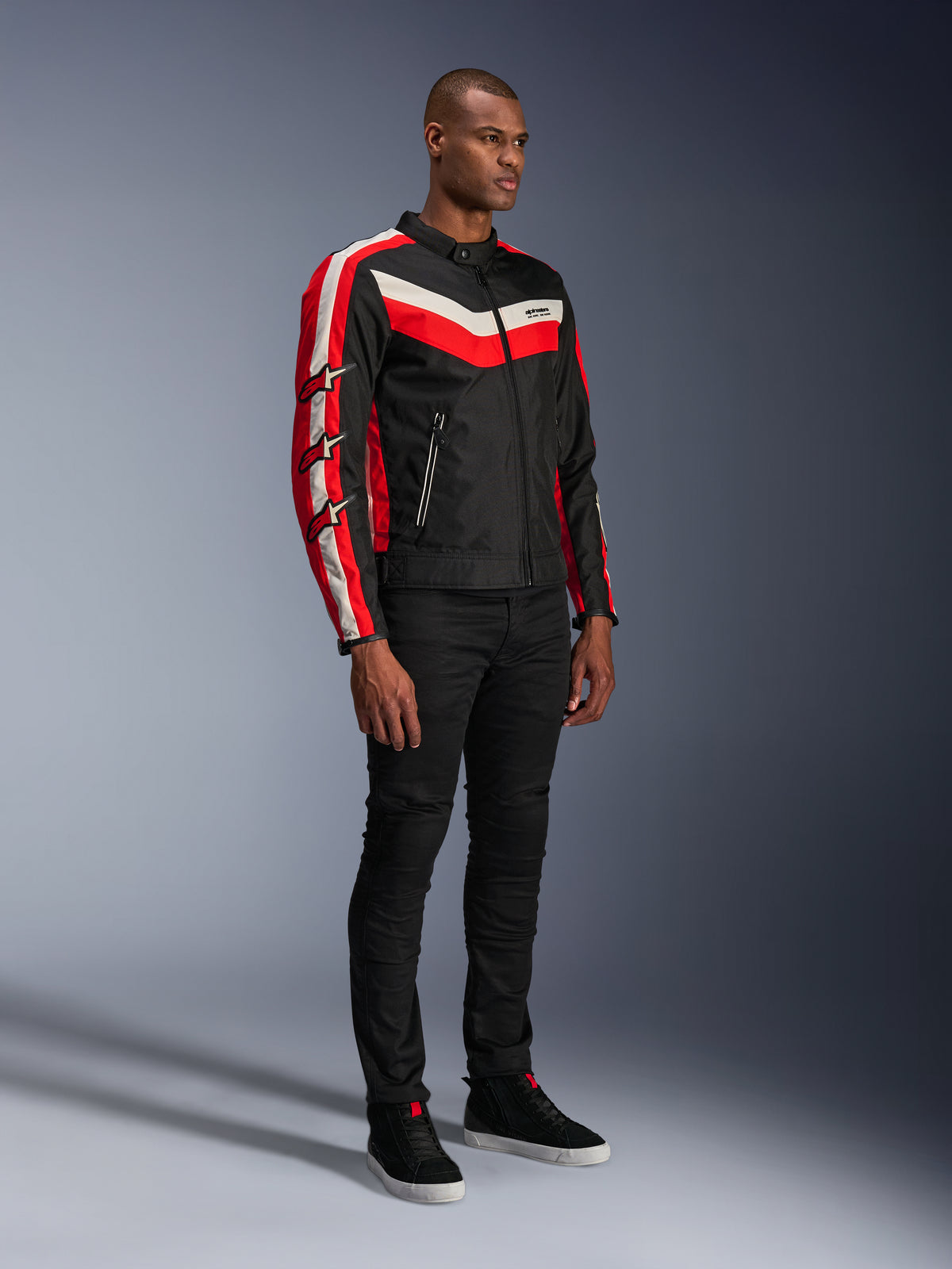 Męski model w kurtce Alpinestars T-Dyno WR, tekstylna kurtka motocyklowa w kolorze Black Bright Red Ecru, czerwone i kremowe pasy, logo gwiazdy Alpinestars na rękawach, czarne spodnie i czarne wysokie trampki, miejska odzież motocyklowa w stylu vintage.