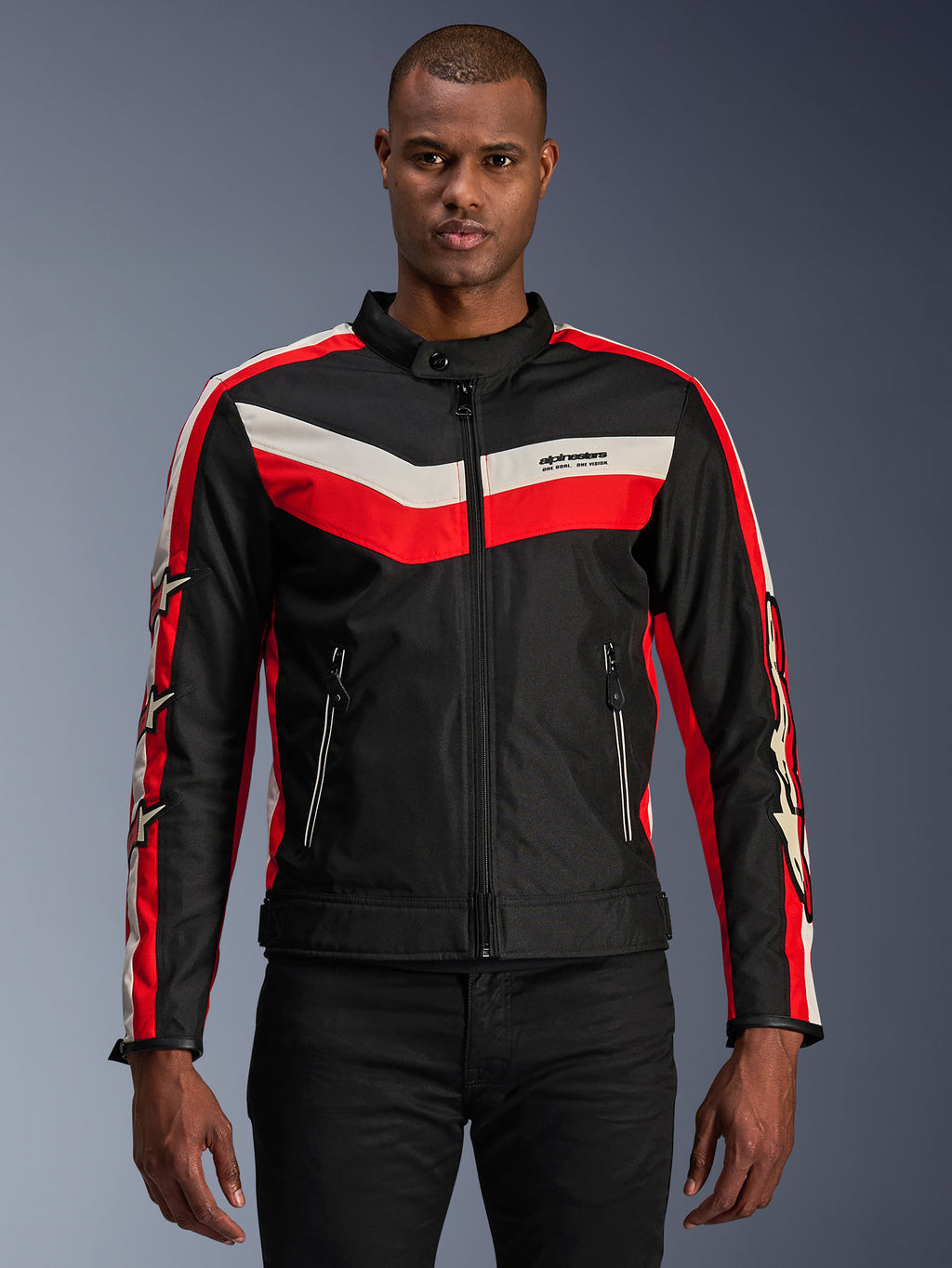 Alpinestars T-Dyno WR Jacket, tekstylna kurtka motocyklowa, czarna z jaskrawoczerwonymi i kremowymi pasami chevron, krój motocyklowy w stylu vintage, wysoki kołnierz z zapięciem na guzik, przednie kieszenie na zamek, logo z gwiazdą na rękawach, noszona przez modela