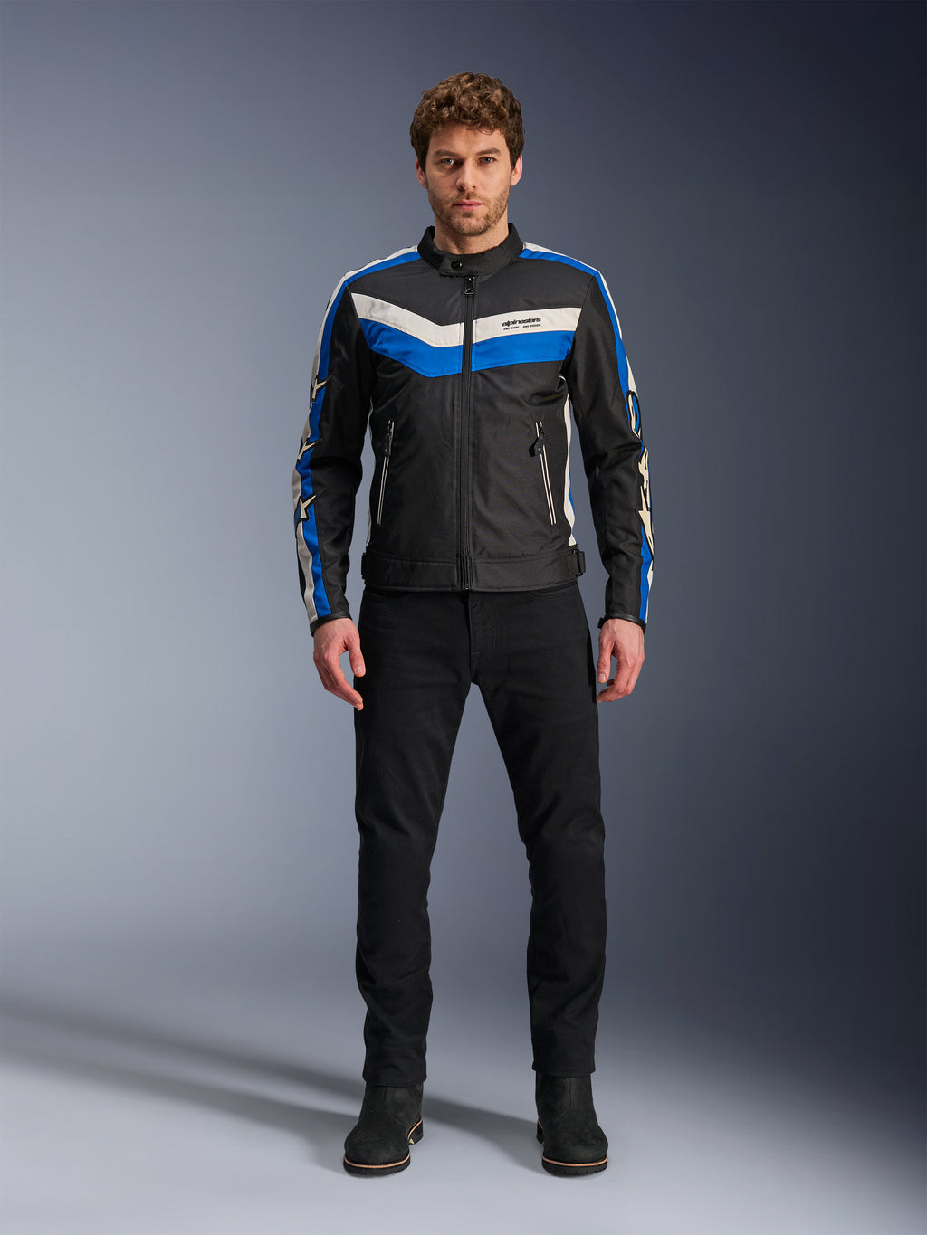 Kurtka Alpinestars T-Dyno WR, tekstylna kurtka motocyklowa, czarna, niebieska i ecru, prezentowana przez modela w czarnych spodniach i butach, z pasami w kształcie szewronu na klatce piersiowej i designem w stylu heritage biker