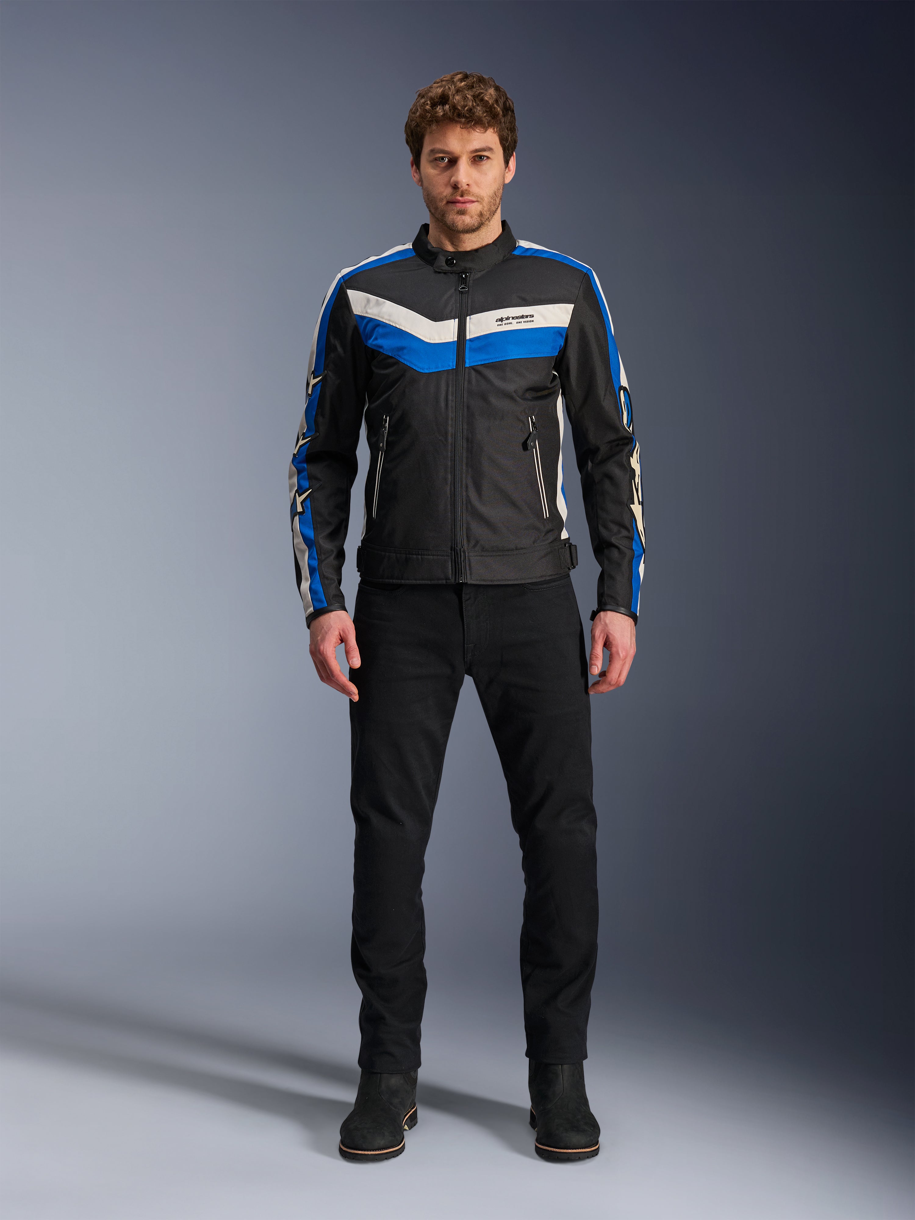 Kurtka Alpinestars T-Dyno WR, tekstylna kurtka motocyklowa, czarna, niebieska i ecru, prezentowana przez modela w czarnych spodniach i butach, z pasami w kształcie szewronu na klatce piersiowej i designem w stylu heritage biker