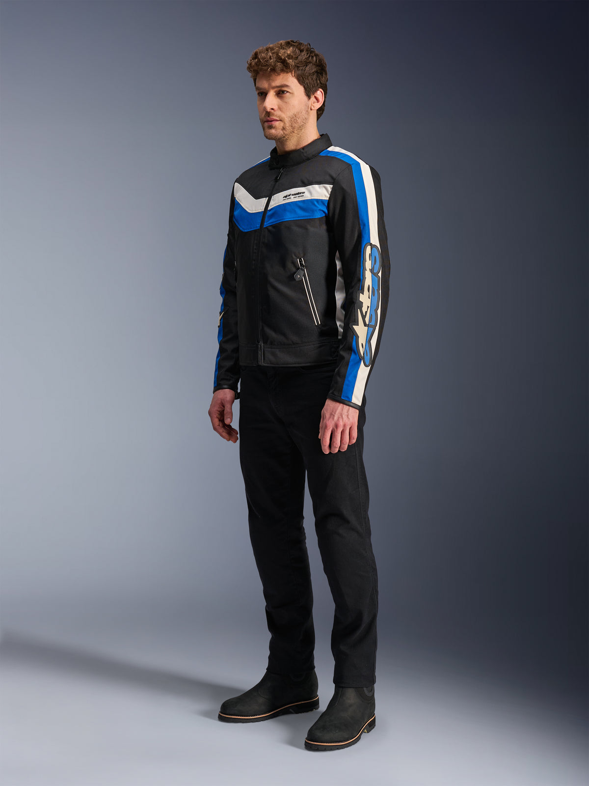 Kurtka Alpinestars T-Dyno WR, czarno-niebieska i ecru, tekstylna kurtka motocyklowa noszona przez modela w czarnych spodniach i butach, design inspirowany stylem vintage z wyraźnymi niebieskimi i kremowymi pasami oraz brandingiem na rękawach, fotografia studyjna