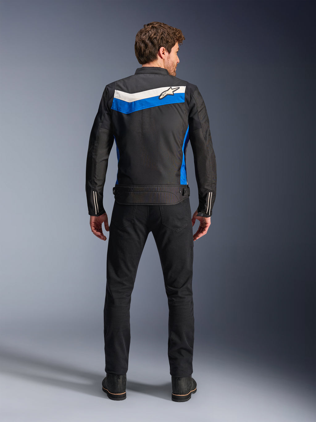 Kurtka Alpinestars T-Dyno WR, tekstylna kurtka motocyklowa, kolorystyka Black Blue Ecru, pełny widok od tyłu na modelu, z niebiesko-kremowym pasem w kształcie szewronu i logo Alpinestars, zestawiona z czarnymi spodniami i butami, miejska odzież motocyklowa inspirowana stylem vintage