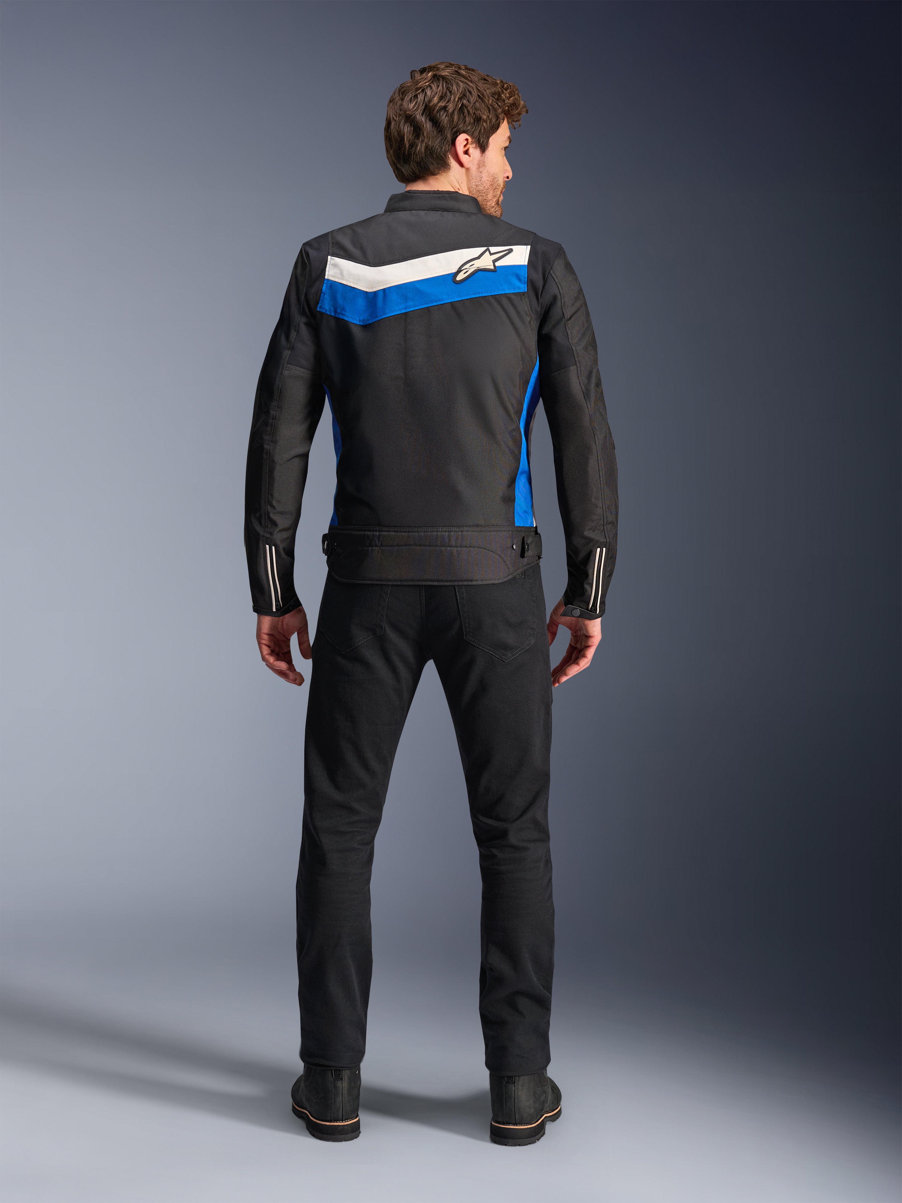 Kurtka Alpinestars T-Dyno WR, tekstylna kurtka motocyklowa, kolorystyka Black Blue Ecru, pełny widok od tyłu na modelu, z niebiesko-kremowym pasem w kształcie szewronu i logo Alpinestars, zestawiona z czarnymi spodniami i butami, miejska odzież motocyklowa inspirowana stylem vintage