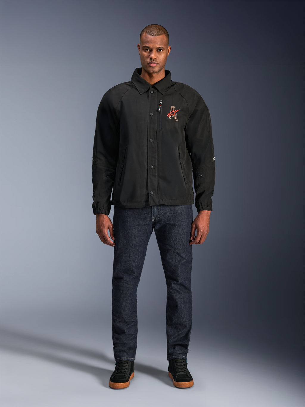 Czarna kurtka Alpinestars Revved WR, model w ciemnoniebieskich dżinsach i czarnych sneakersach, widok z przodu, zapięcie na zatrzaski i haftowane logo 3D na piersi, współczesna miejska odzież motocyklowa