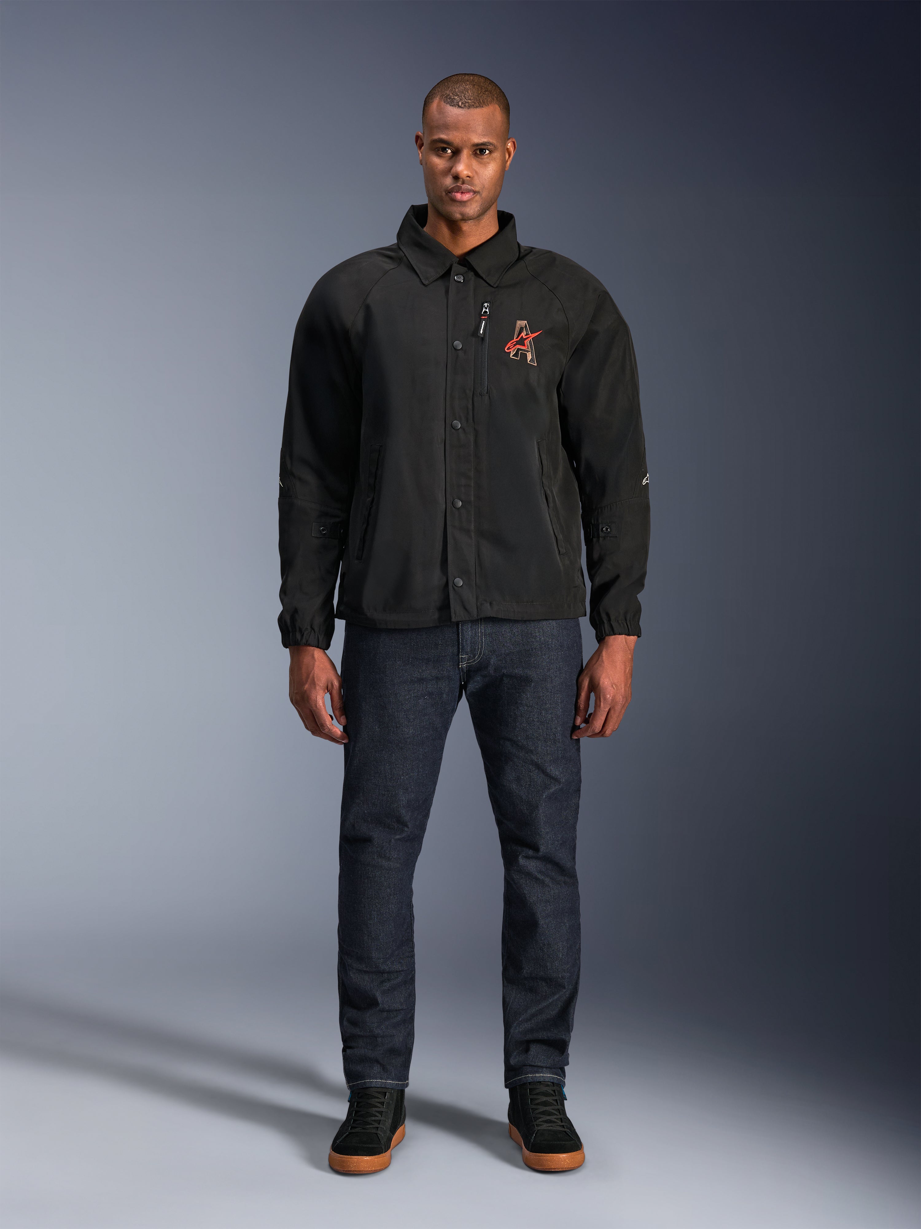 Czarna kurtka Alpinestars Revved WR, model w ciemnoniebieskich dżinsach i czarnych sneakersach, widok z przodu, zapięcie na zatrzaski i haftowane logo 3D na piersi, współczesna miejska odzież motocyklowa