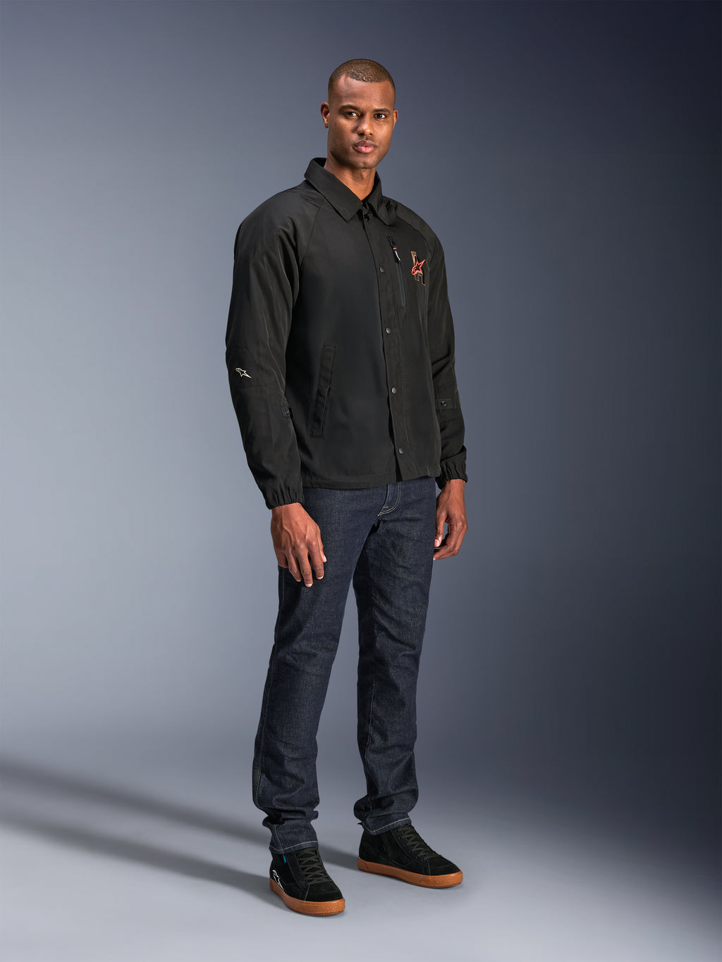 Alpinestars Revved WR Jacket w kolorze czarnym, miejska kurtka motocyklowa w stylu trenerskim noszona przez modela z ciemnoniebieskimi dżinsami i czarnymi sneakersami, z zapięciem na zatrzaski i haftowanym logo, minimalistyczny i sportowy design do jazdy szosowej.