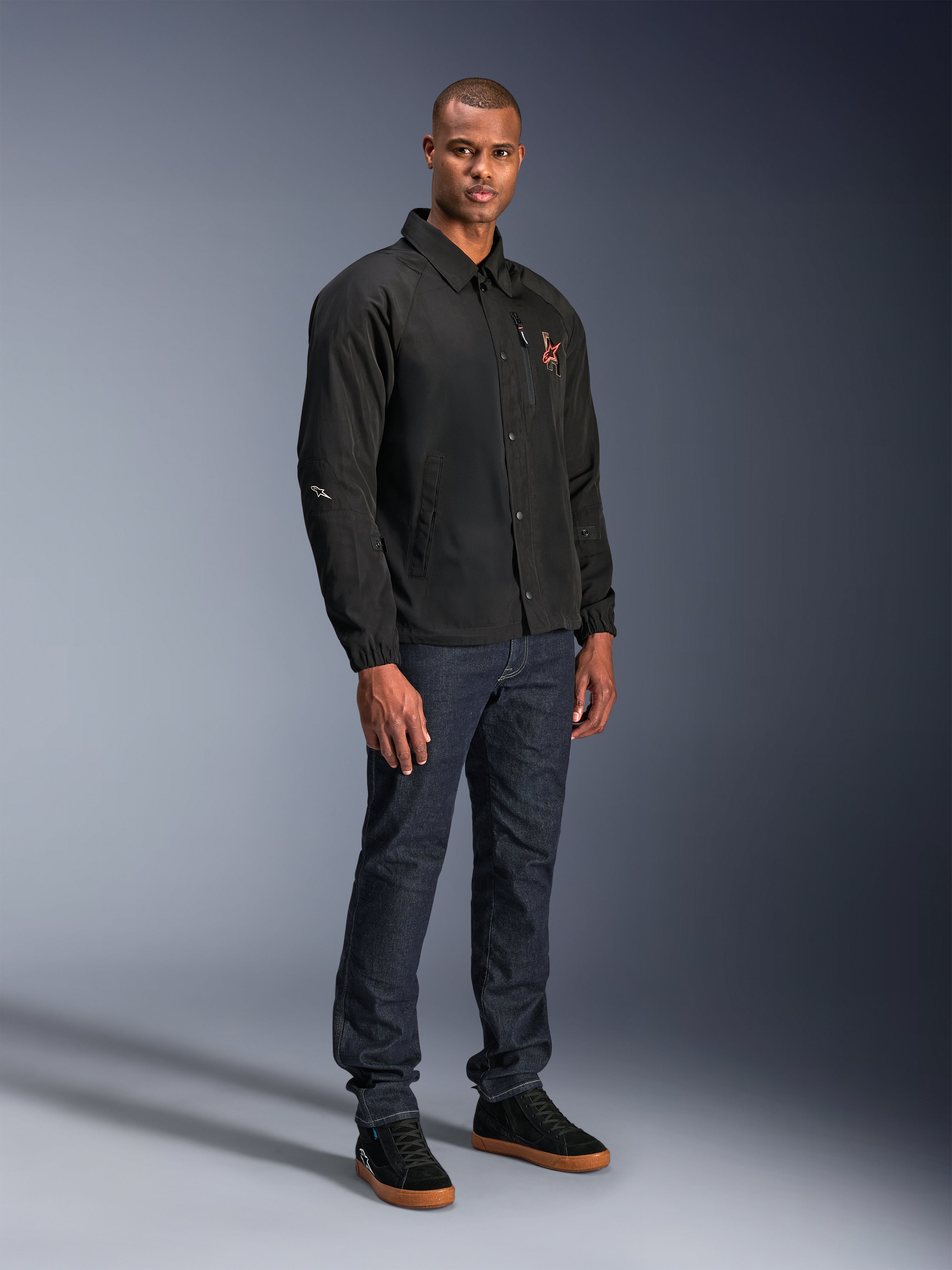 Alpinestars Revved WR Jacket w kolorze czarnym, miejska kurtka motocyklowa w stylu trenerskim noszona przez modela z ciemnoniebieskimi dżinsami i czarnymi sneakersami, z zapięciem na zatrzaski i haftowanym logo, minimalistyczny i sportowy design do jazdy szosowej.