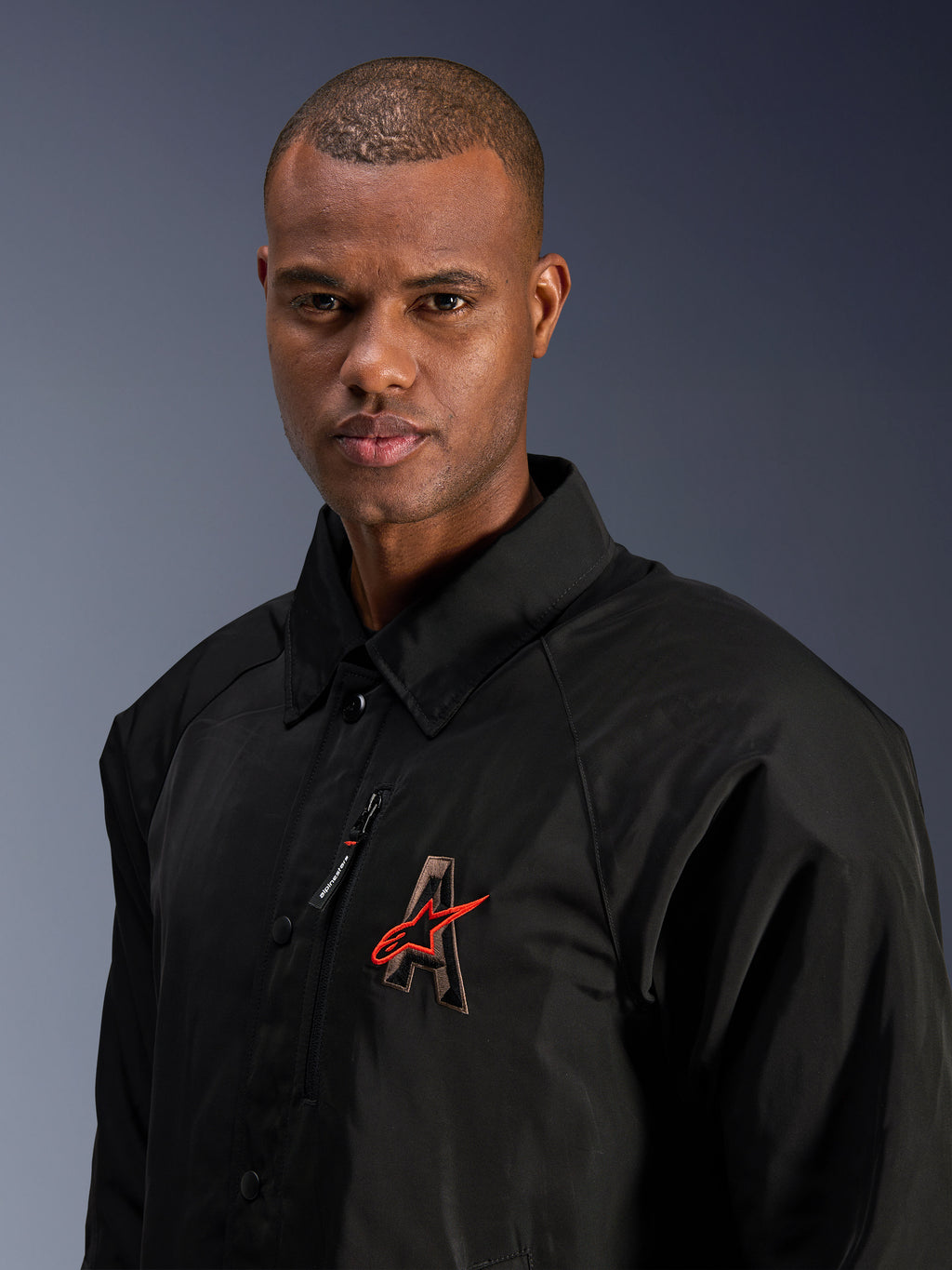Alpinestars Revved WR Jacket, czarna, zbliżenie na mężczyznę w wodoodpornej satynowej kurtce motocyklowej, haftowane logo „A” 3D z czerwoną gwiazdą Alpinestars, kieszeń na piersi na zamek, kołnierzyk, nowoczesny styl miejski