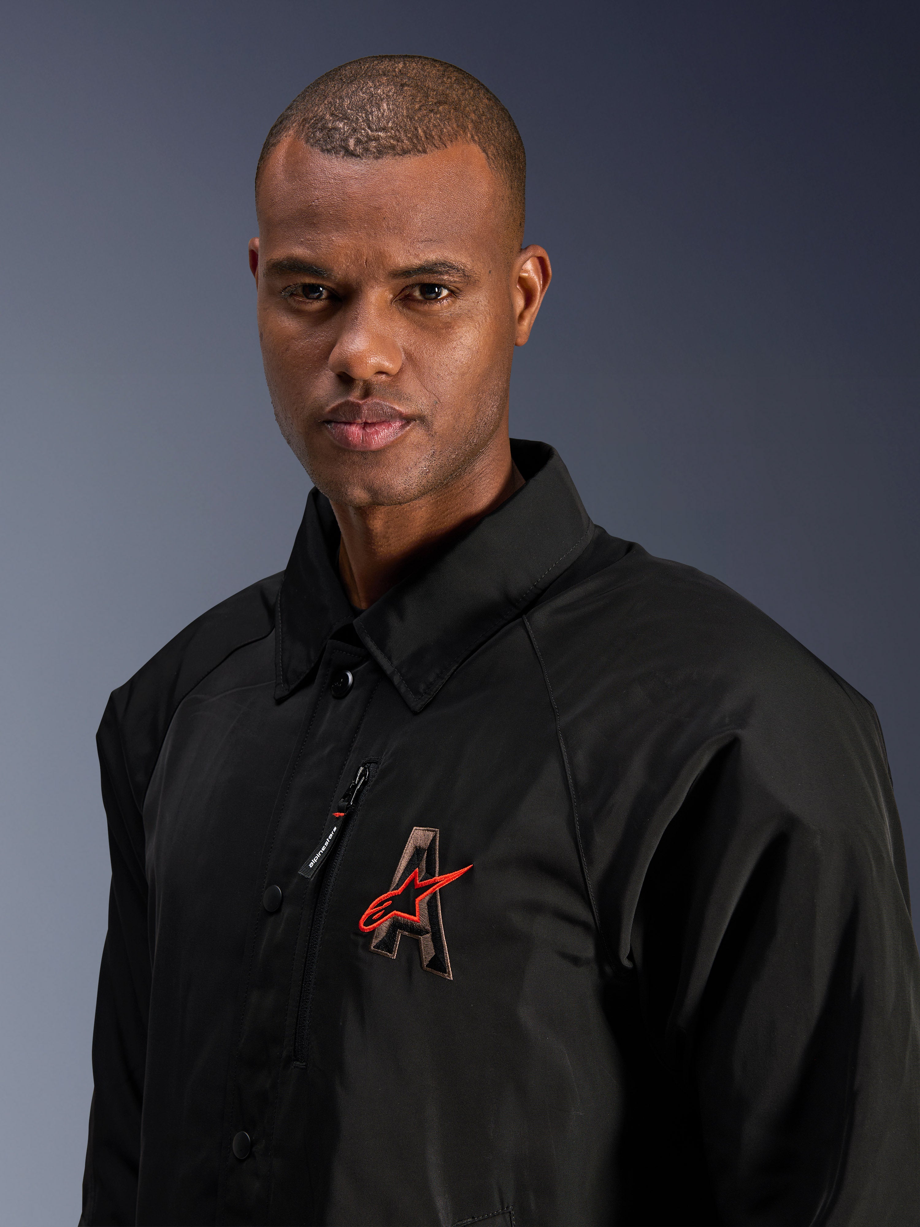 Alpinestars Revved WR Jacket, czarna, zbliżenie na mężczyznę w wodoodpornej satynowej kurtce motocyklowej, haftowane logo „A” 3D z czerwoną gwiazdą Alpinestars, kieszeń na piersi na zamek, kołnierzyk, nowoczesny styl miejski