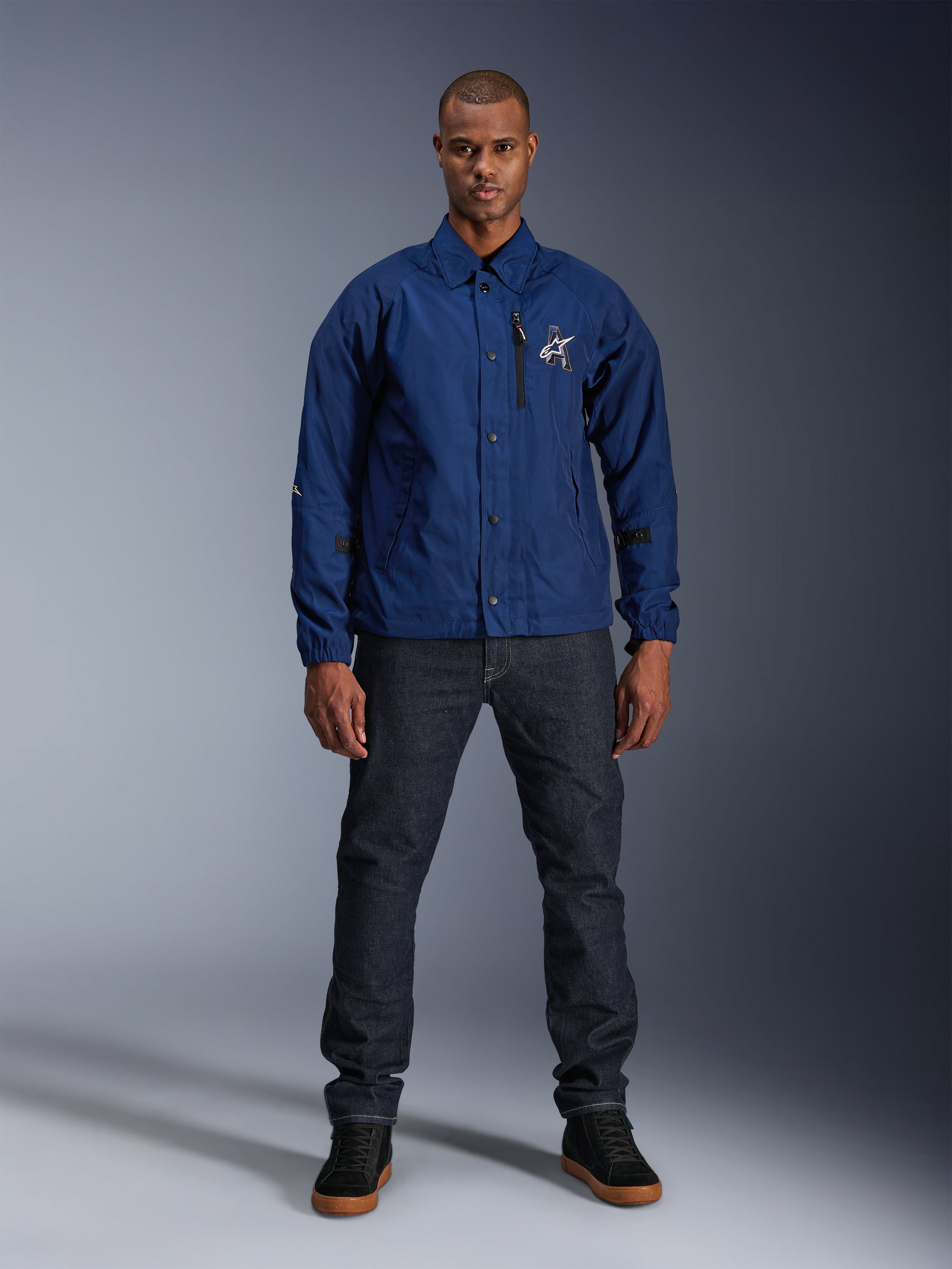 Kurtka Alpinestars Revved WR, granatowa, motocyklowa kurtka w stylu trenerskim, noszona przez mężczyznę w ciemnych jeansach i czarnych sneakersach, z zapięciem na zatrzaski i logo Alpinestars 3D, współczesny miejski styl streetwear