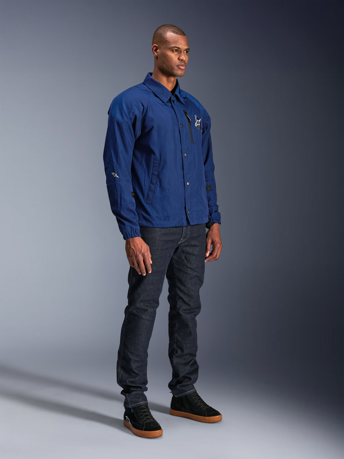 Kurtka Alpinestars Revved WR, niebieska kurtka motocyklowa, noszona przez modela w ciemnych jeansach i czarnych sneakersach, wodoodporna sylwetka typu coaches z zapięciem na zatrzaski i minimalistycznym logo na piersi.
